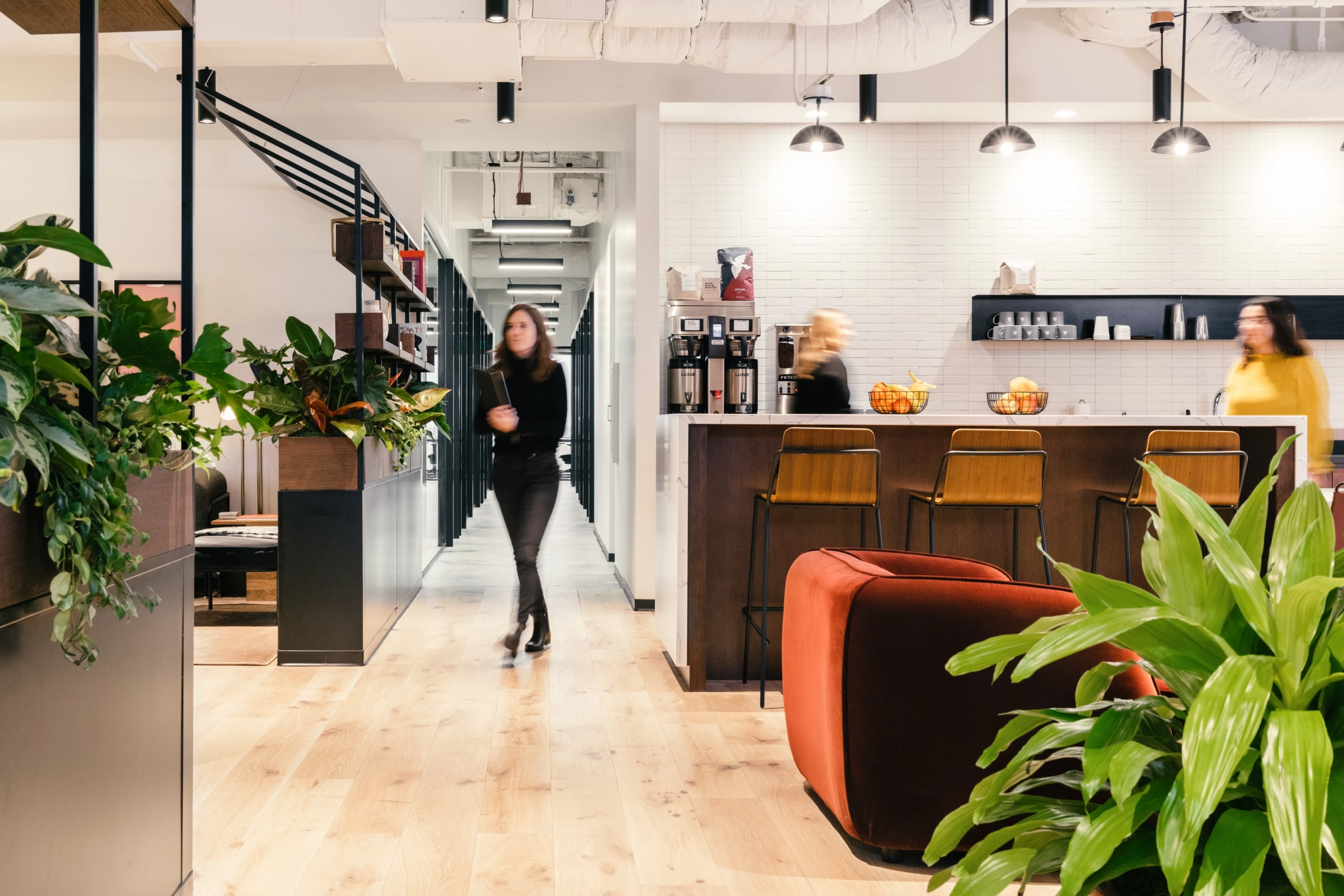 Moderne coworkingruimte met een koffiebar, planten en wandelende mensen. De inrichting bestaat uit houten vloeren, hanglampen en een bar met krukken, waardoor een uitnodigende sfeer van samenwerking ontstaat.