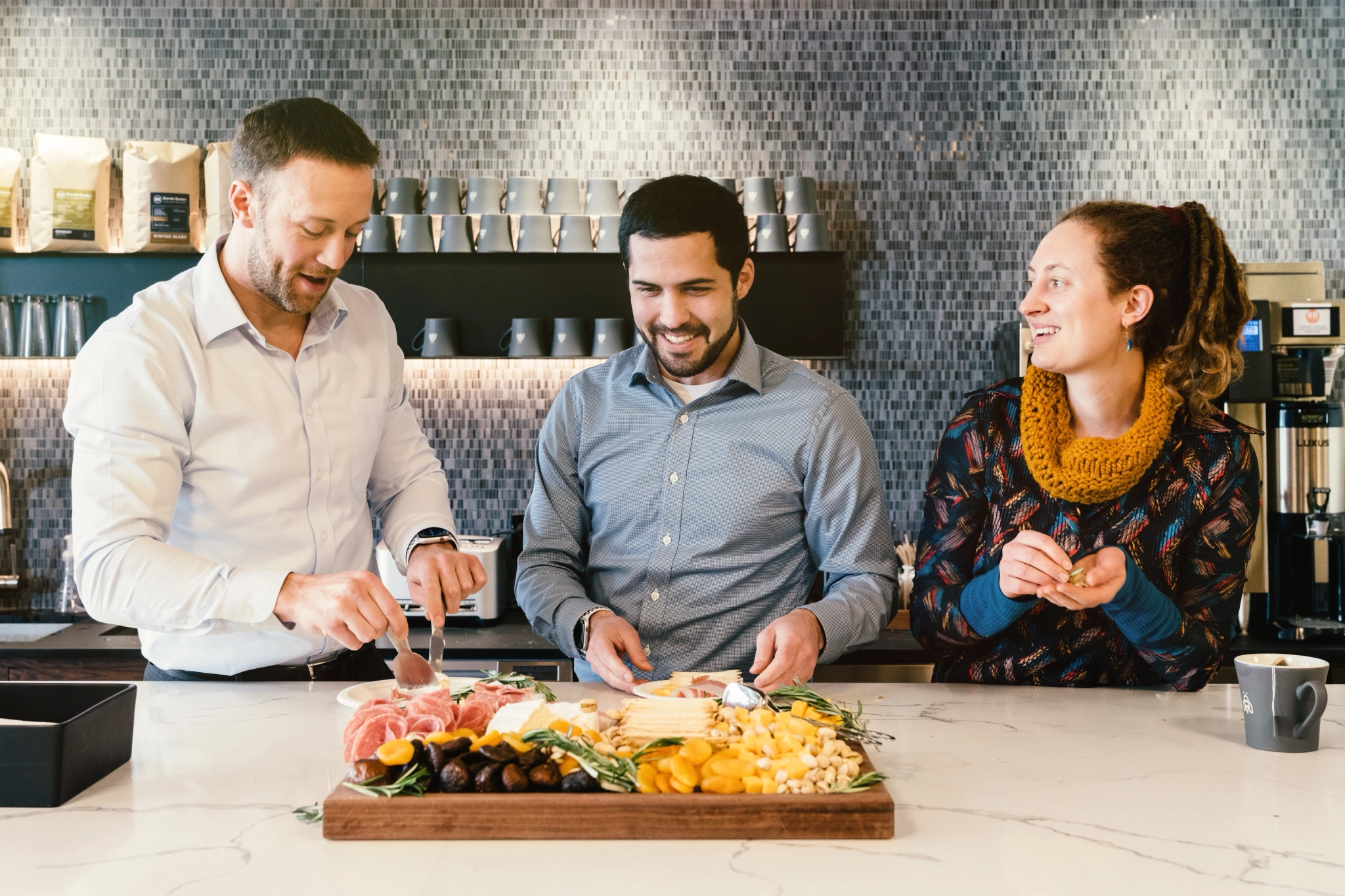 Drei Personen stehen an einer Theke in einem Büro in Portland und bereiten ein Wurstbrett mit verschiedenen Fleischsorten, Käsesorten, Nüssen und Früchten zu und genießen es.