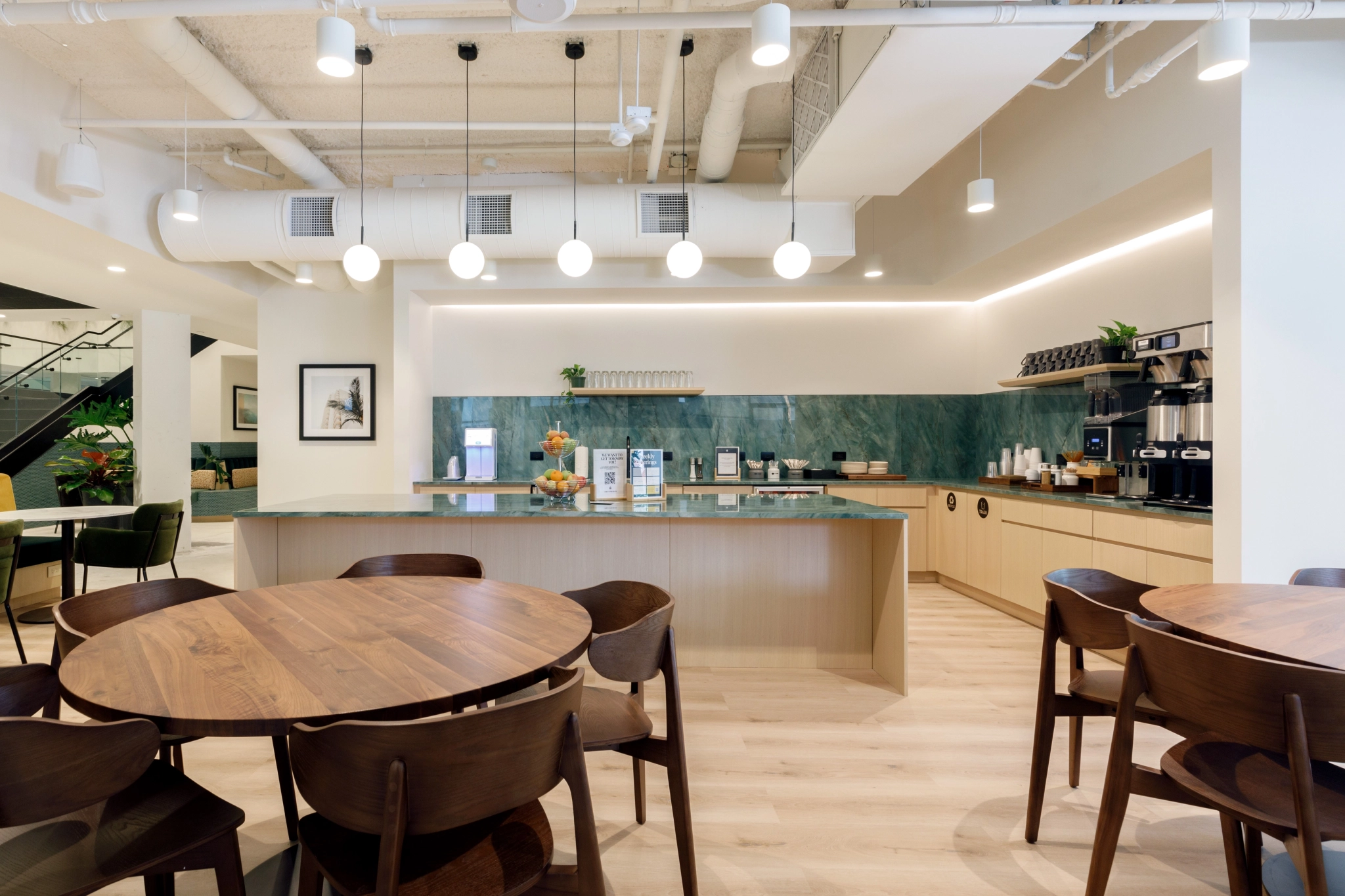 Modern coworking kitchen with wooden tables and chairs, a long counter featuring a marble backsplash, and stylish hanging lights. Coffee machines and a fruit bowl adorn the counter, creating an inviting space for casual conversations or quick breaks from the meeting room.