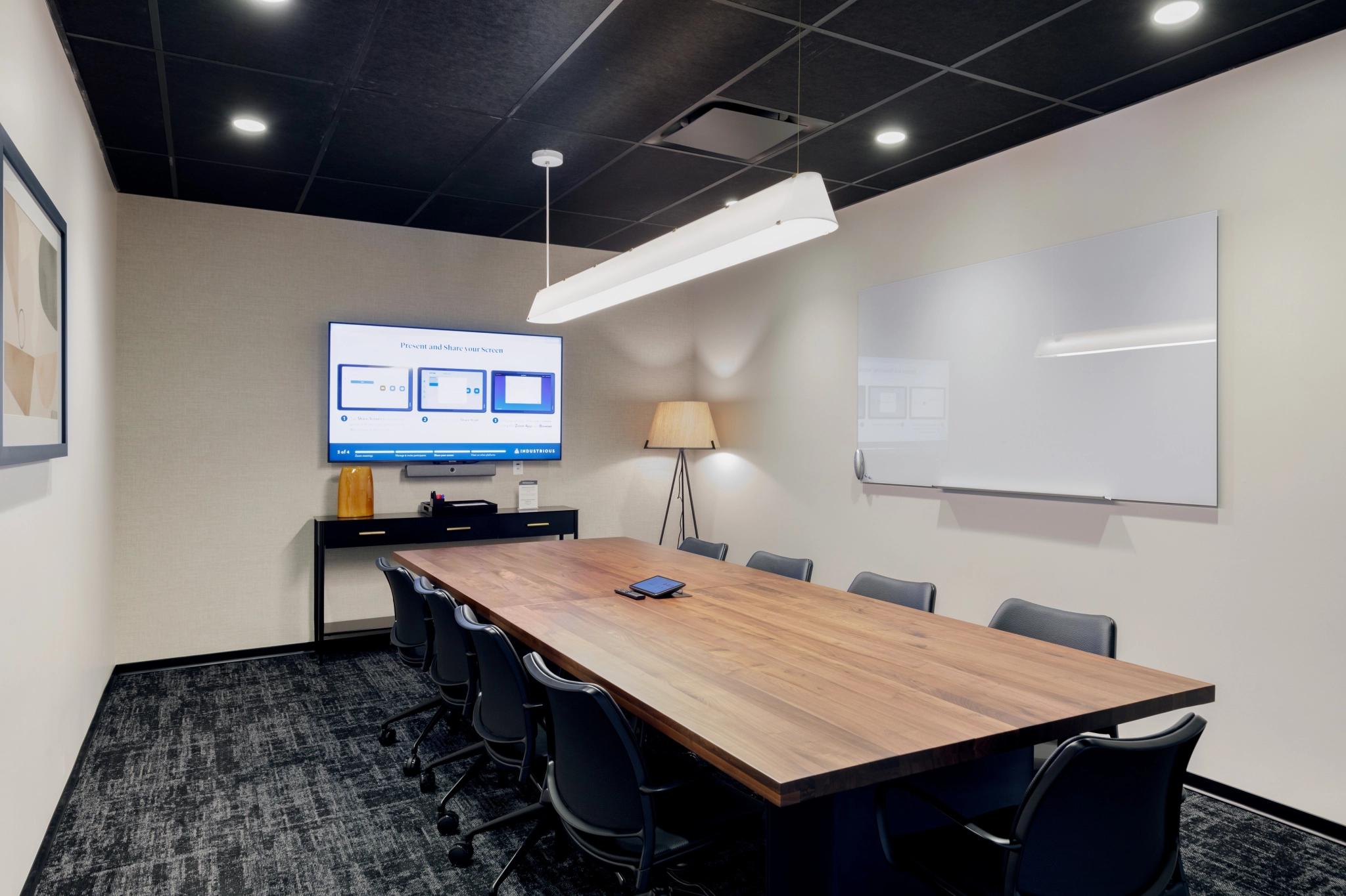 A modern meeting room in Coconut Grove featuring a wooden table, black chairs, and a wall-mounted screen displaying slides. A whiteboard adorns one wall, while a floor lamp and ceiling lights provide ample illumination.