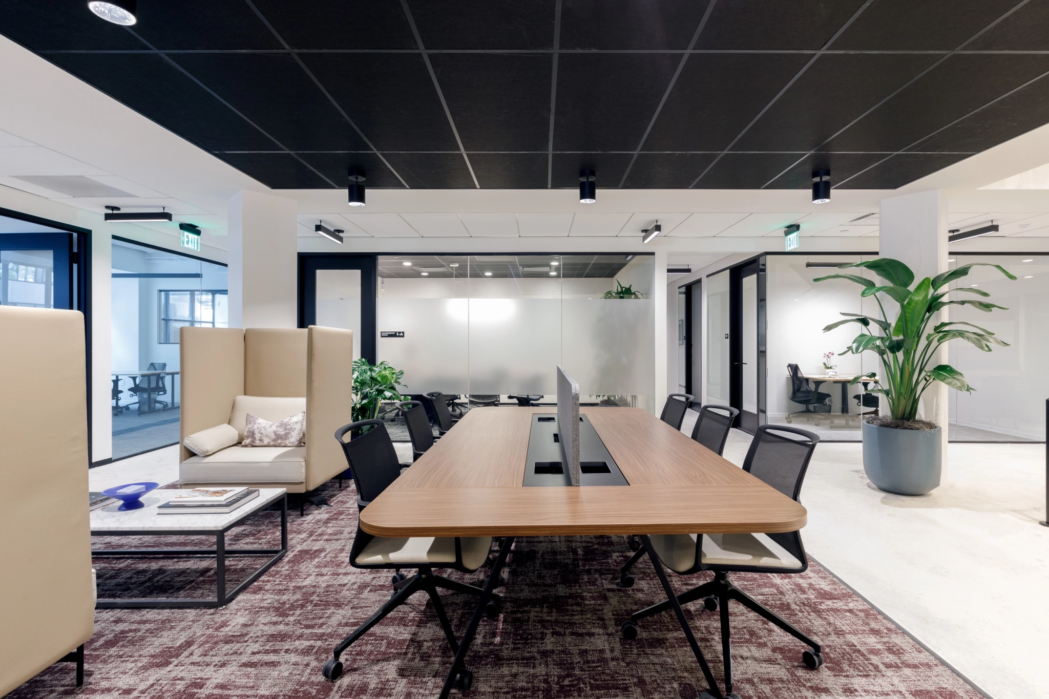 Nestled in Coconut Grove, this modern workspace features a long wooden table, black chairs, and lush plants. Glass partitions elegantly divide the area, with a cozy seating nook nearby. Black ceiling tiles create a striking contrast against the pristine white walls.