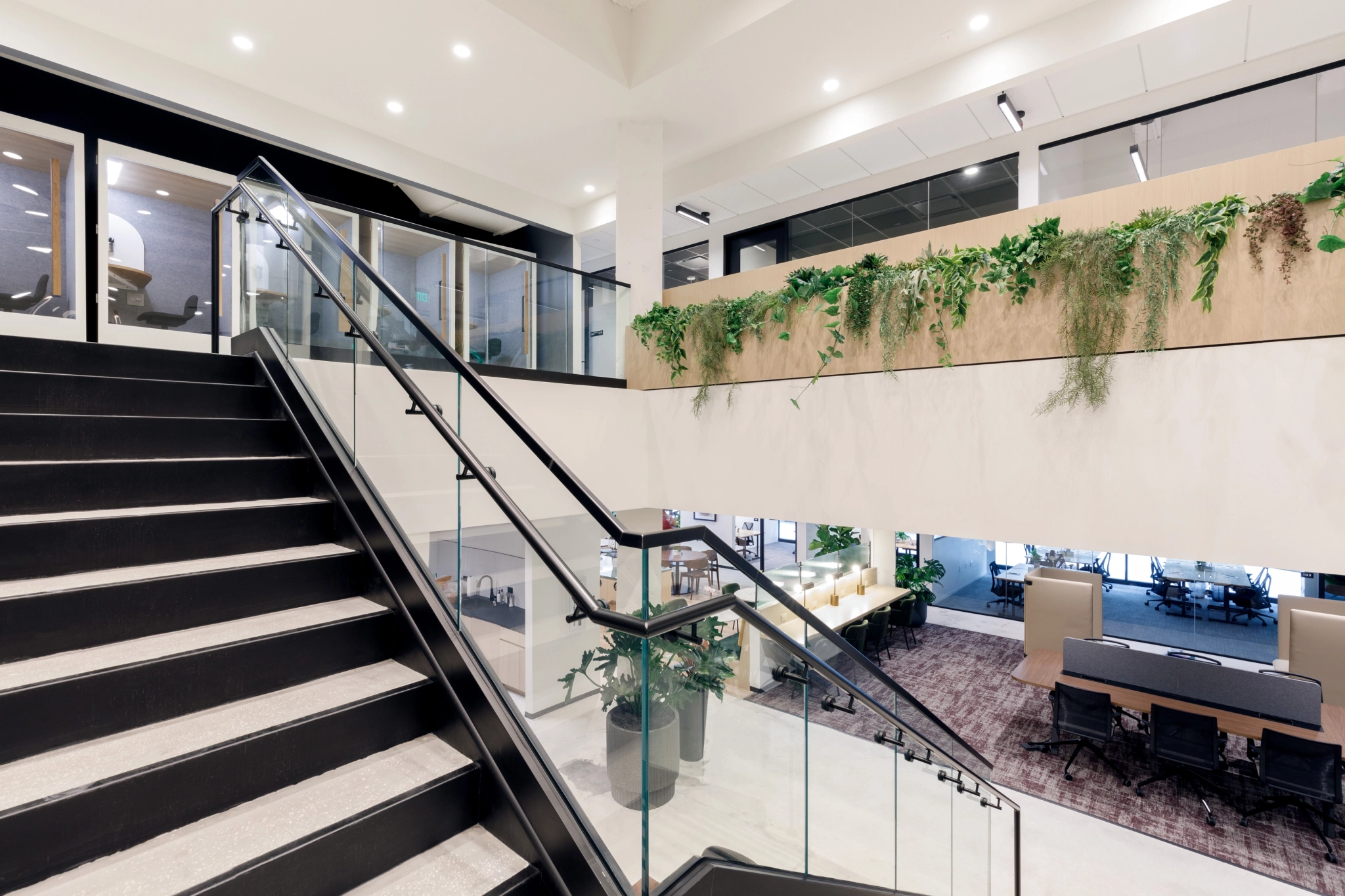 Modern Coconut Grove office interior with glass railings, staircase, and greenery on the upper level. Large open space with desks and a sleek meeting room below. Bright lighting and clean design enhance the vibrant atmosphere.