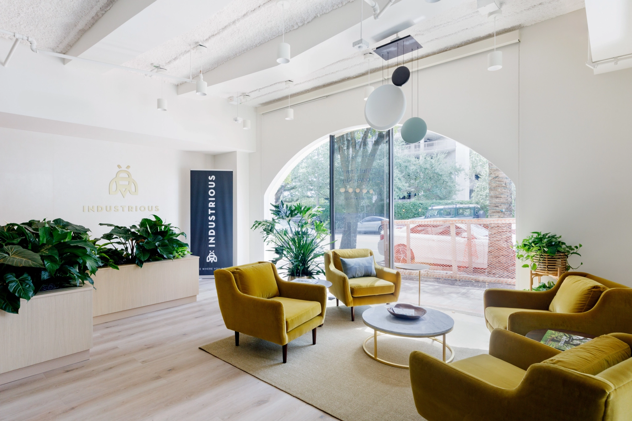 The modern office lobby, located in Coconut Grove, features yellow chairs, green plants, and large windows that flood the coworking space with natural light. A sign reads "Industrious," while a parked orange car is visible outside.