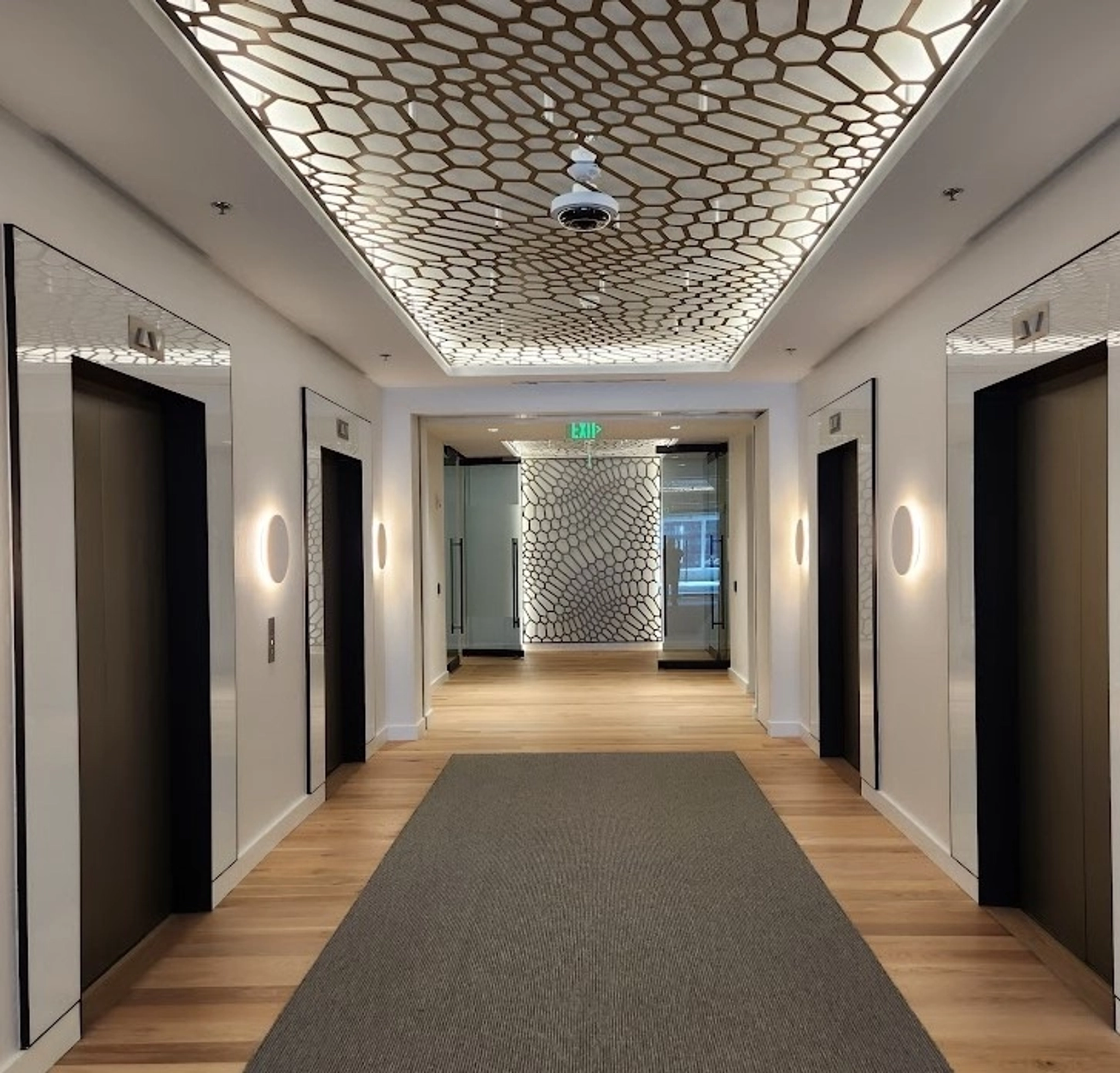 Contemporary office lobby with four elevators, two on each side. The hallway, leading to a modern meeting room and workspace, features a geometric patterned ceiling and wall, wooden flooring, and a grey carpeted area in the center.