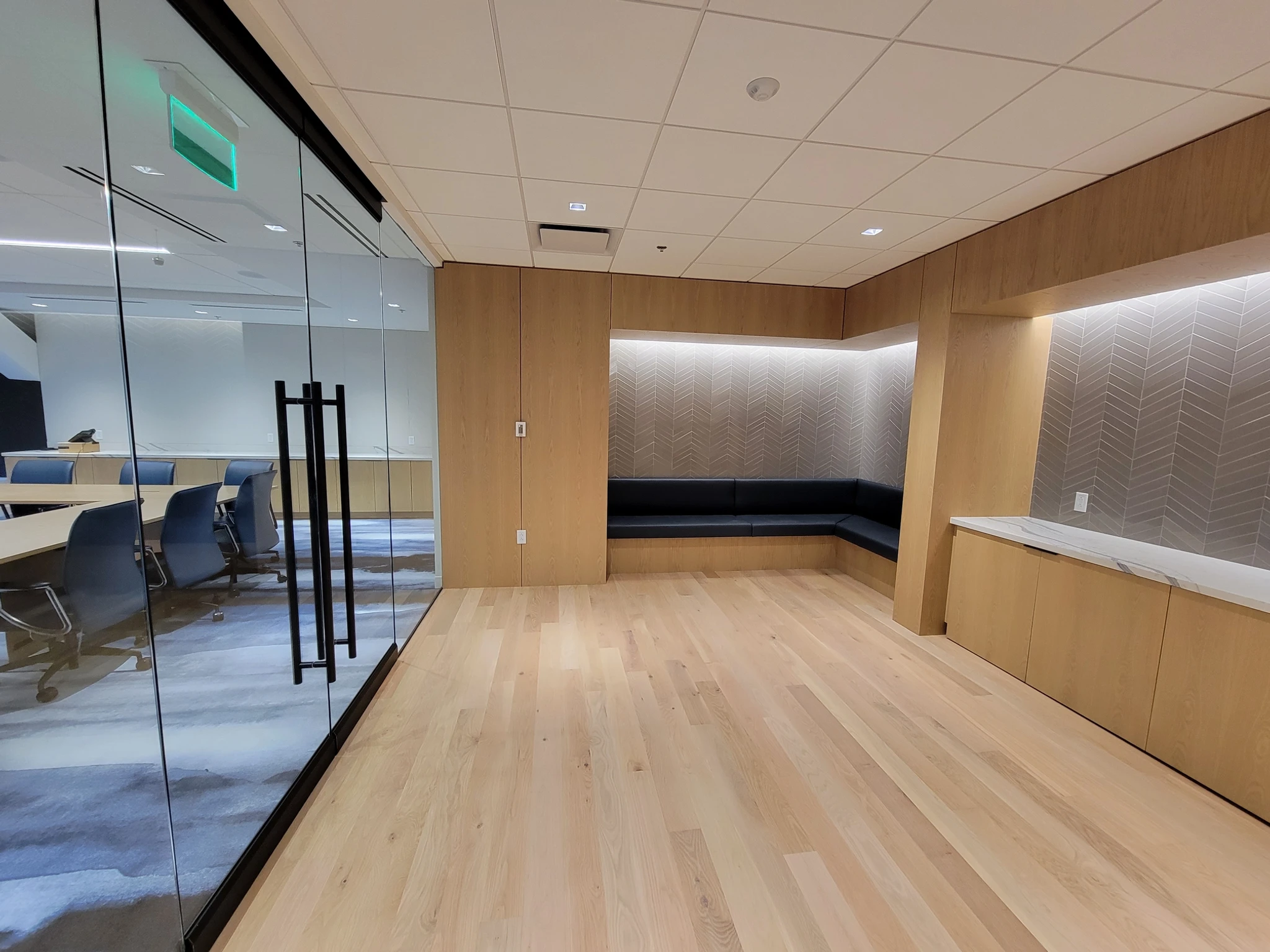 A modern Boston office space featuring wood flooring, recessed seating, and a glass-walled conference room with blue chairs around a large table. Ceiling lights and an exit sign are also visible in this contemporary workspace.