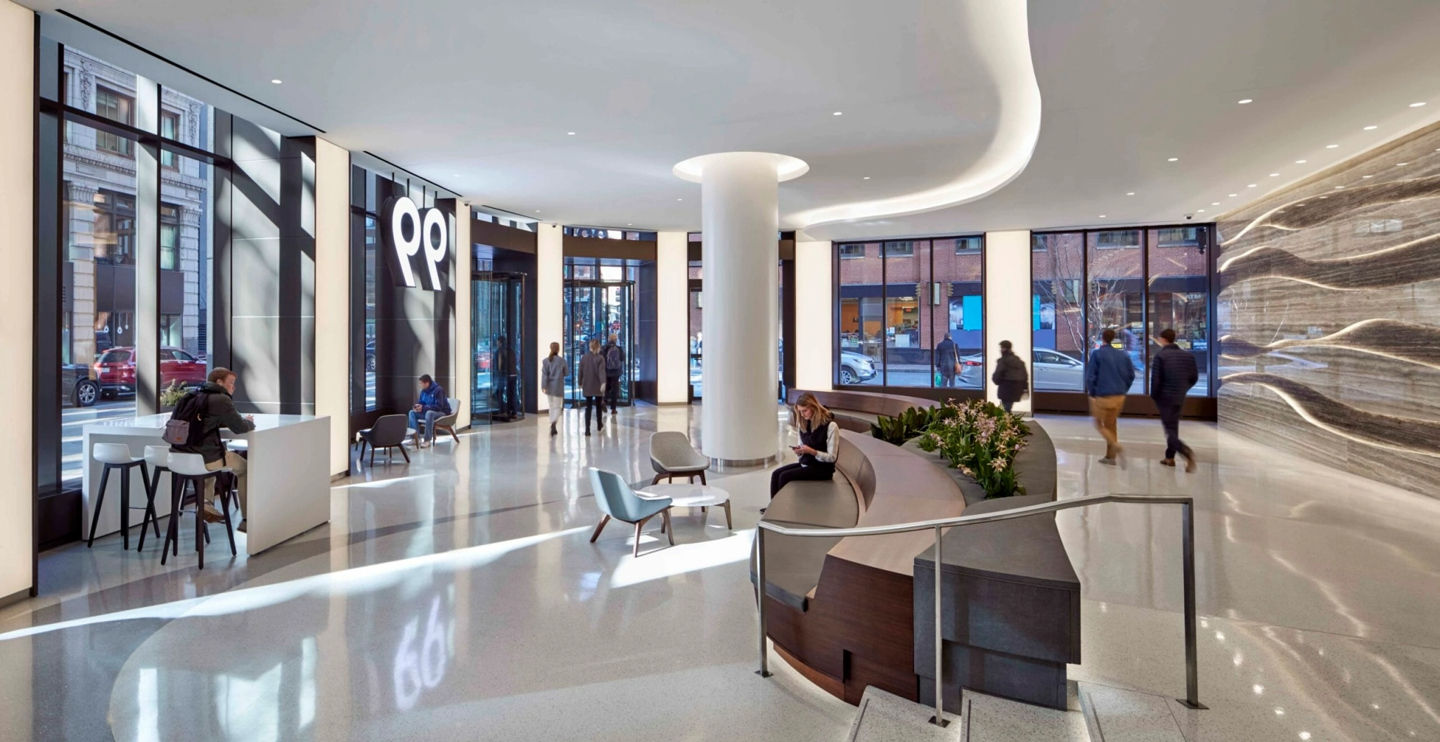 Modern lobby area with large windows, seating arrangements, and people walking and sitting. A white pillar stands in the center, and a curved design element adorns the right wall. Nearby, a glass-enclosed meeting room provides a sleek workspace for professionals.