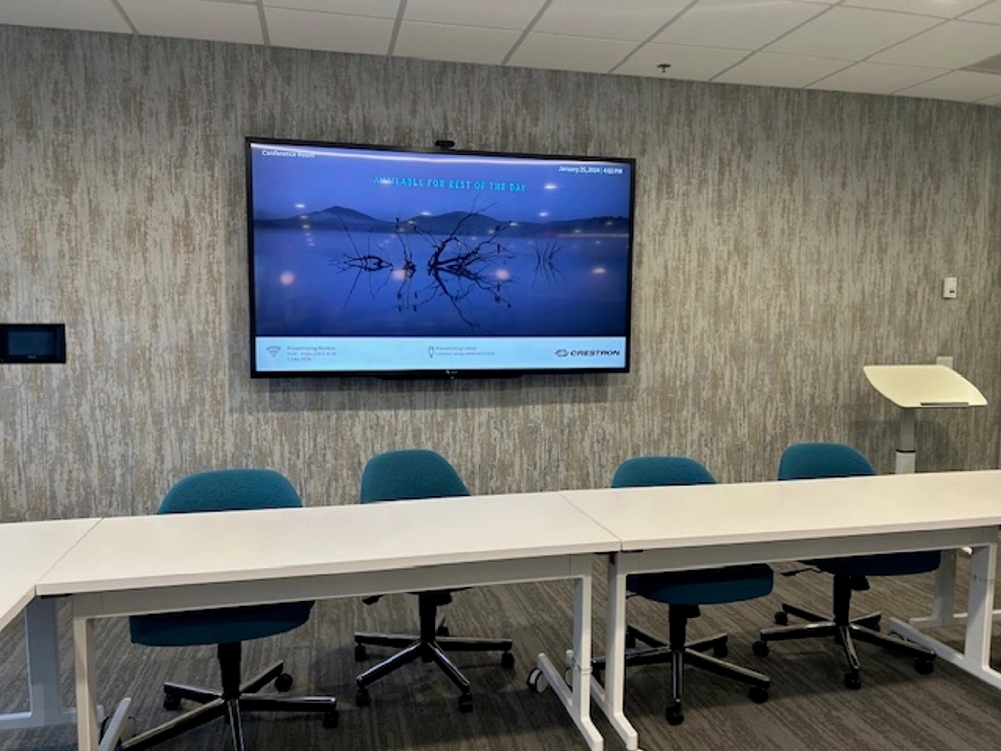 A modern office meeting room with a wall-mounted display screen showing a presentation. Blue office chairs and ceiling vents are visible in this well-designed workspace, featuring a gray textured wall and white ceiling tiles.