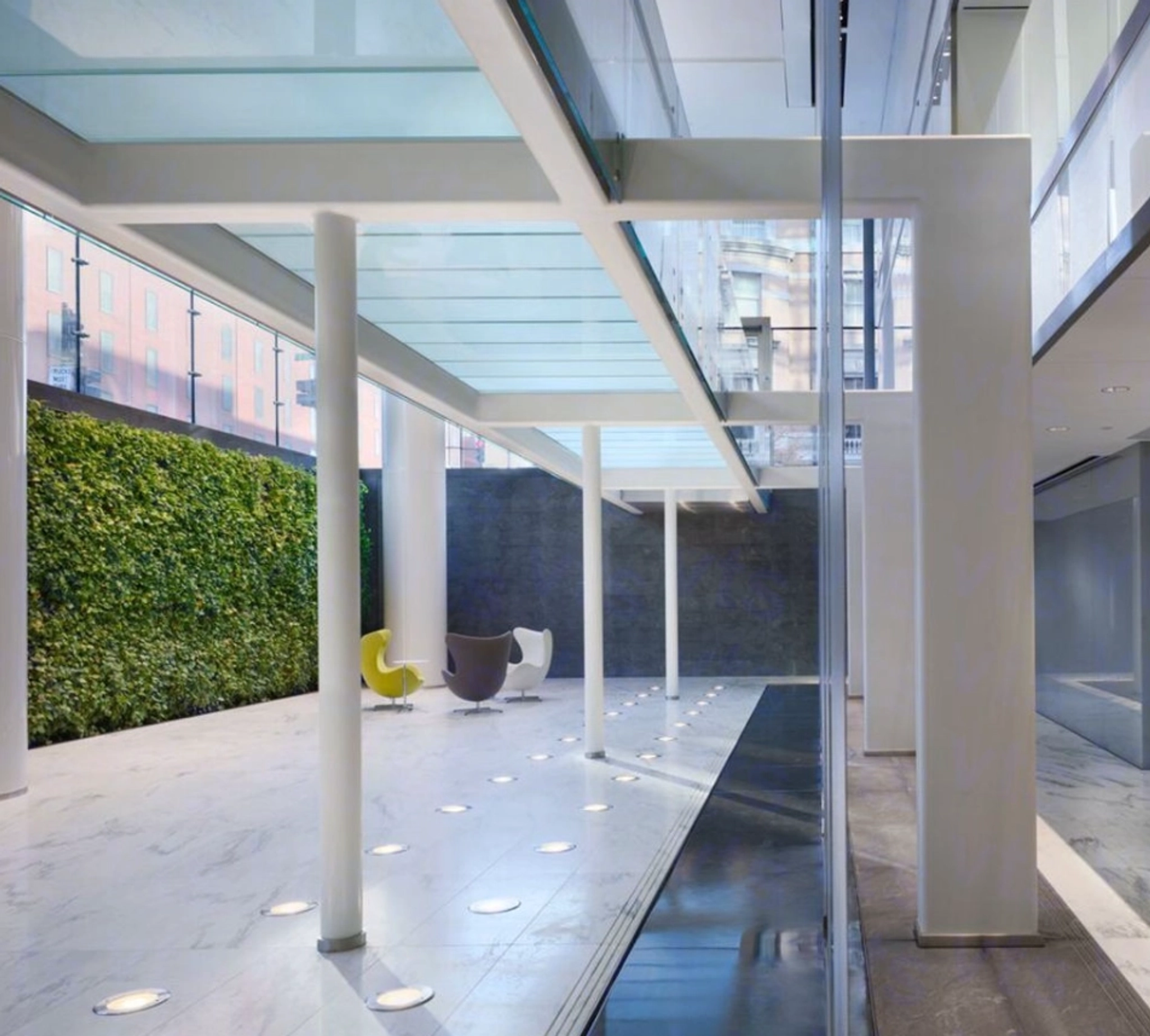 Modern indoor courtyard with a vertical garden, glass ceiling, and minimalist seating. White pillars support the structure, and recessed lighting dots the marble floor. Perfect for an office or coworking space looking to combine nature with productivity.