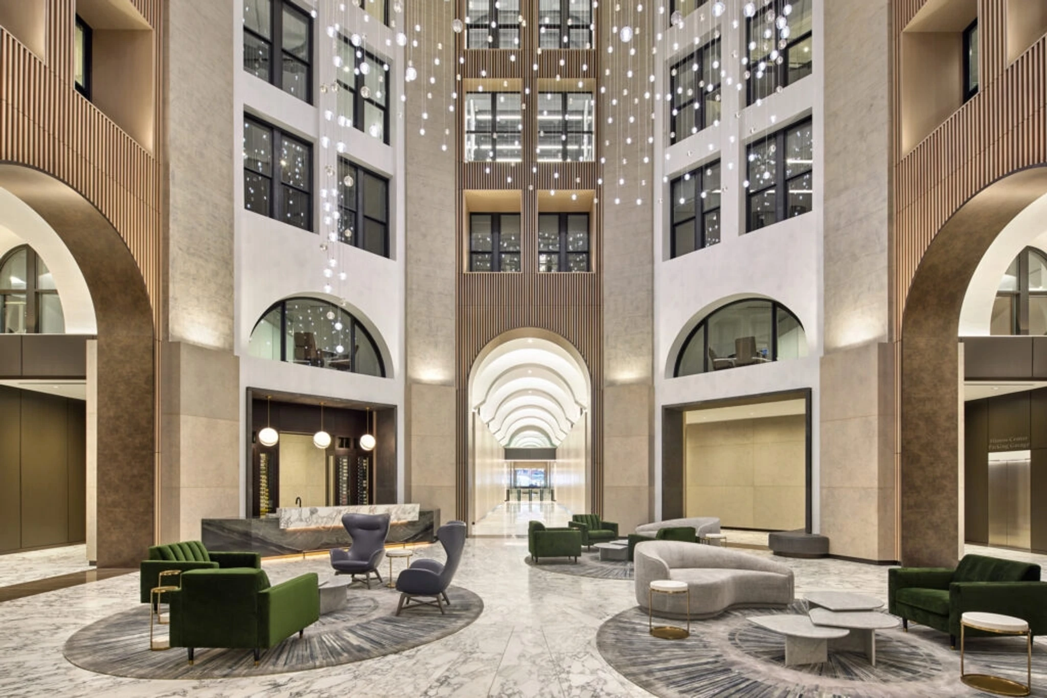 The image shows a modern, spacious hotel lobby with tall ceilings, contemporary furniture, and a large chandelier. The room features a mix of gray and green seating areas around circular rugs, creating an inviting atmosphere that seamlessly doubles as a coworking space.