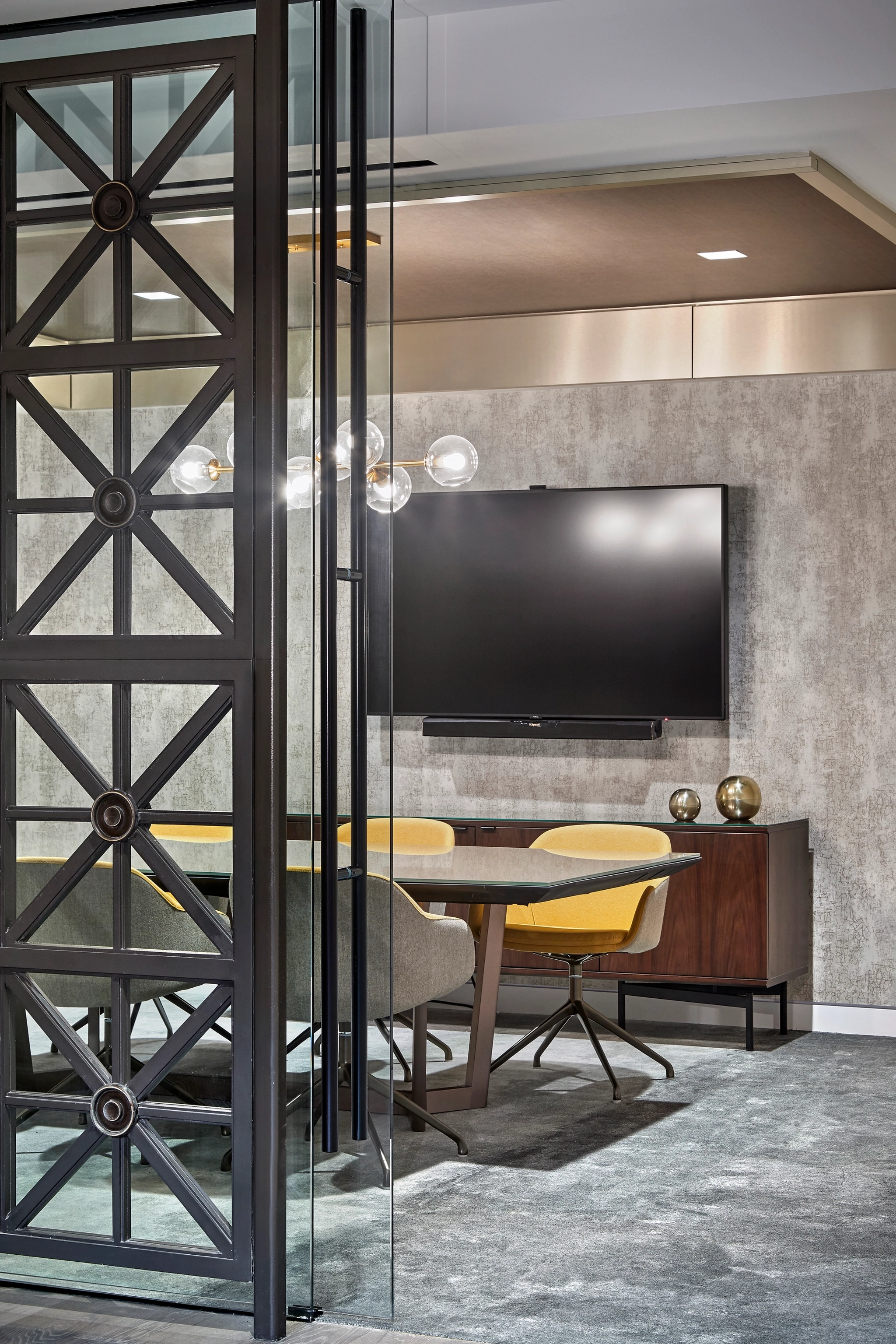 A modern Washington meeting room featuring a large wall-mounted TV, a wooden cabinet, a round table surrounded by yellow and gray chairs, and a glass partition with a metal frame.