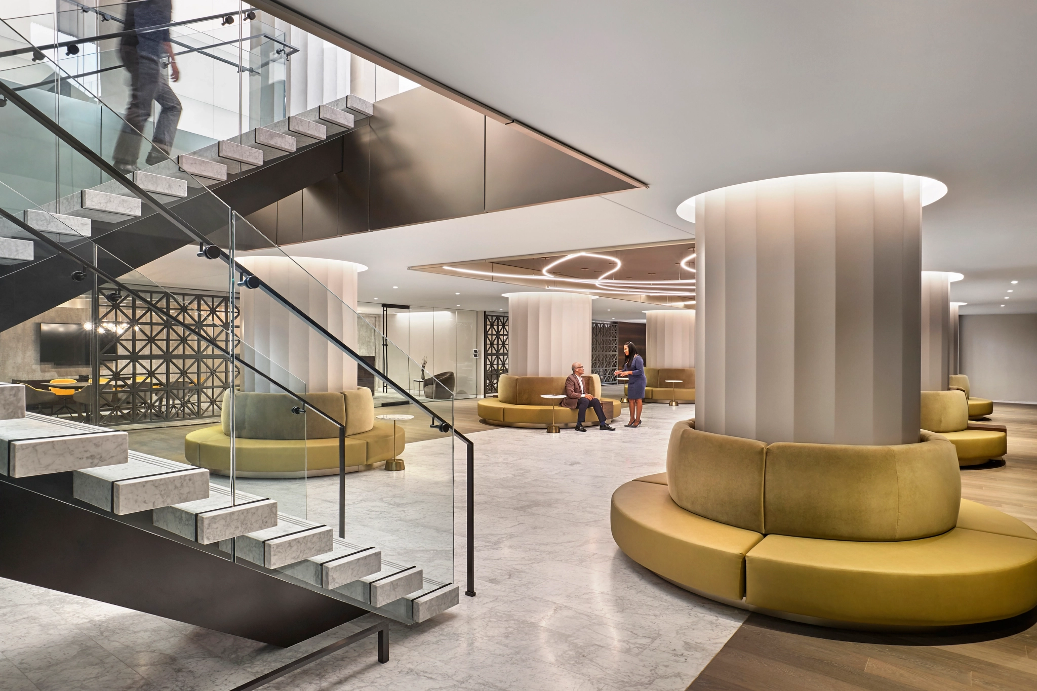 Spacious modern lobby with a large staircase, plush circular seating, and two people conversing. Neutral tones and soft lighting enhance the contemporary design, perfect for a coworking workspace in WASHINGTON.