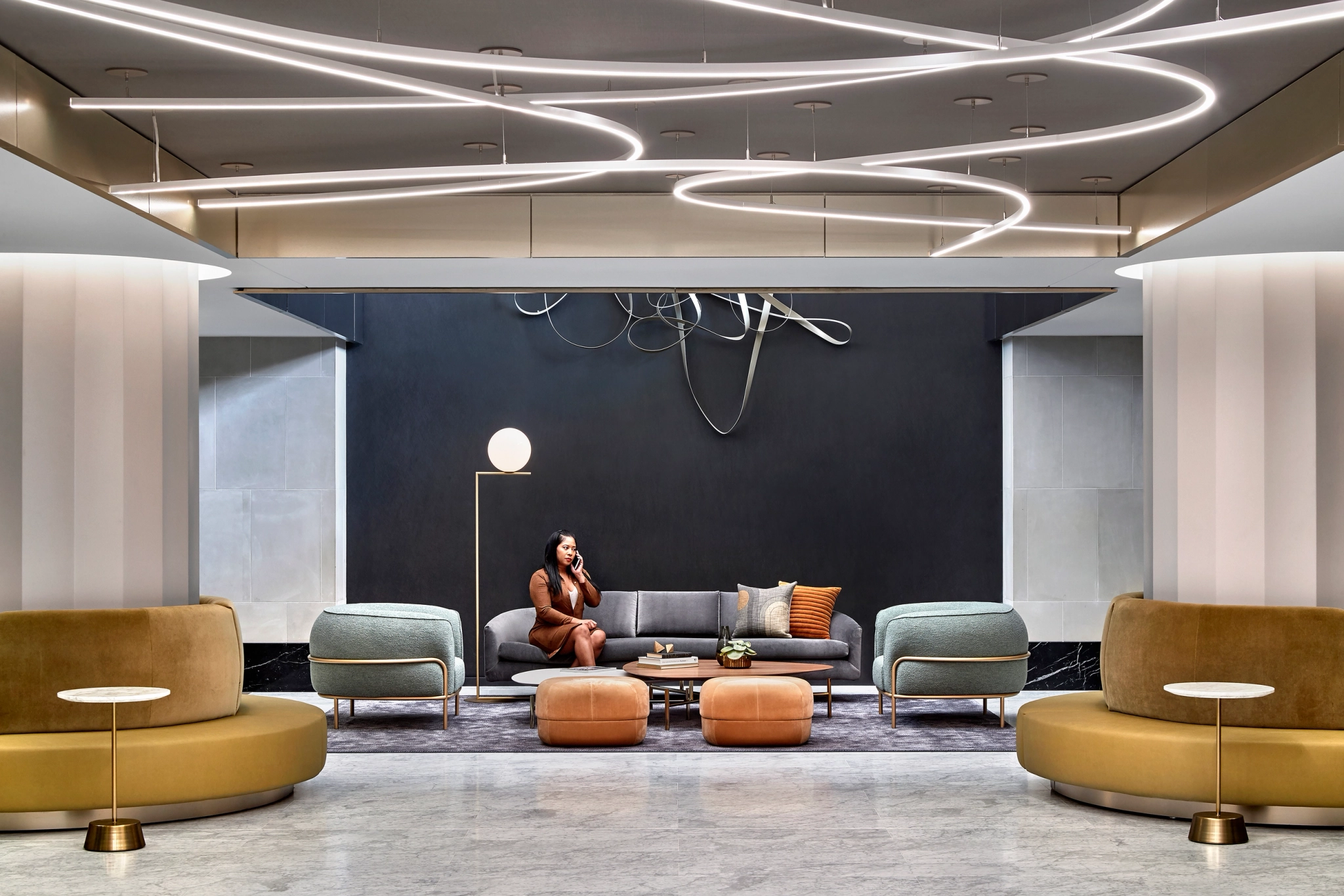 A woman on the phone sits on a modern couch in a sleek coworking lobby with minimalist furniture and abstract lighting fixtures on the ceiling.