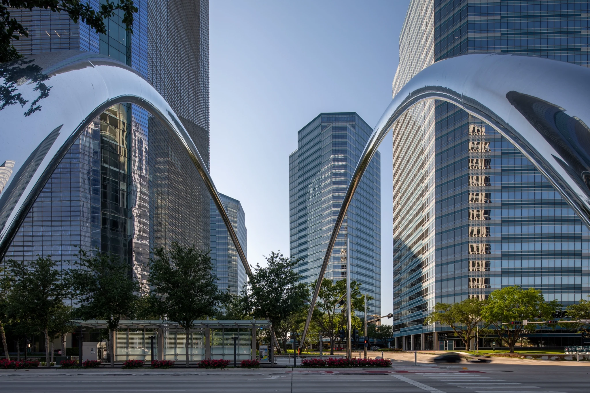 Modern cityscape featuring tall glass buildings under a clear sky with large metallic arches and lush greenery in the foreground, seamlessly blending into innovative coworking spaces.