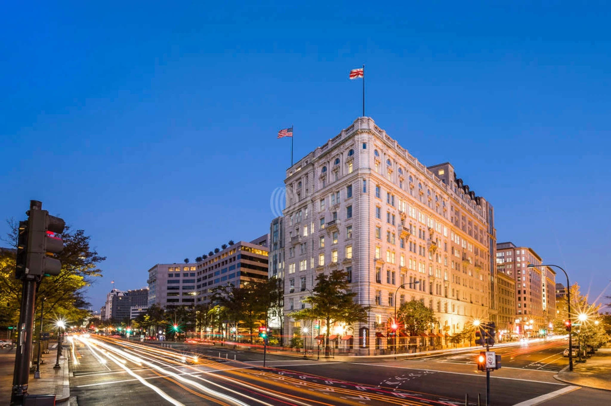 Evening view of a busy city intersection in WASHINGTON with light trails from vehicles, adjacent to a large multi-story building with an American flag on top, near a bustling coworking space.