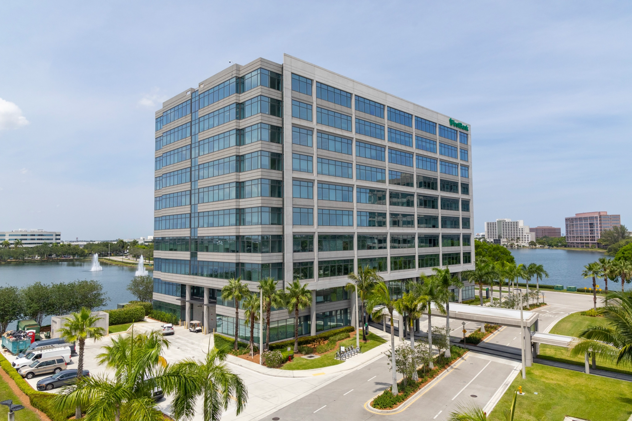 Een modern kantoorgebouw van glas en staal met meerdere verdiepingen in Miami wordt omgeven door palmbomen met een waterkant op de achtergrond. Op het wateroppervlak is een fontein zichtbaar, waardoor een ideale coworking-ruimte ontstaat te midden van een prachtig uitzicht.