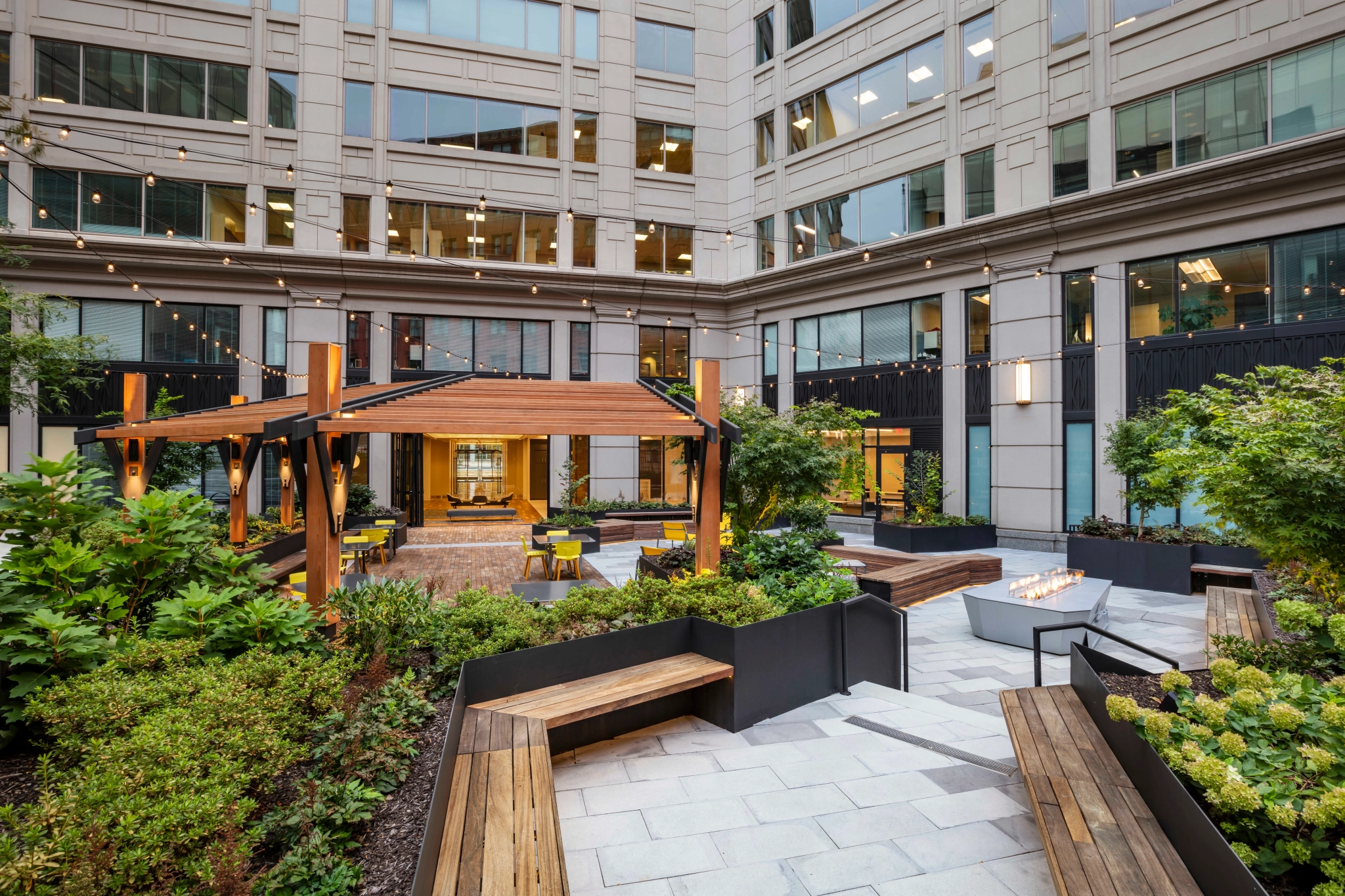 Outdoor courtyard with a wooden pergola, benches, greenery, and string lights in the heart of a modern Washington office building. Planter boxes teem with various plants and shrubs beside a lit decorative fire feature, creating an ideal gathering space outside your meeting room.