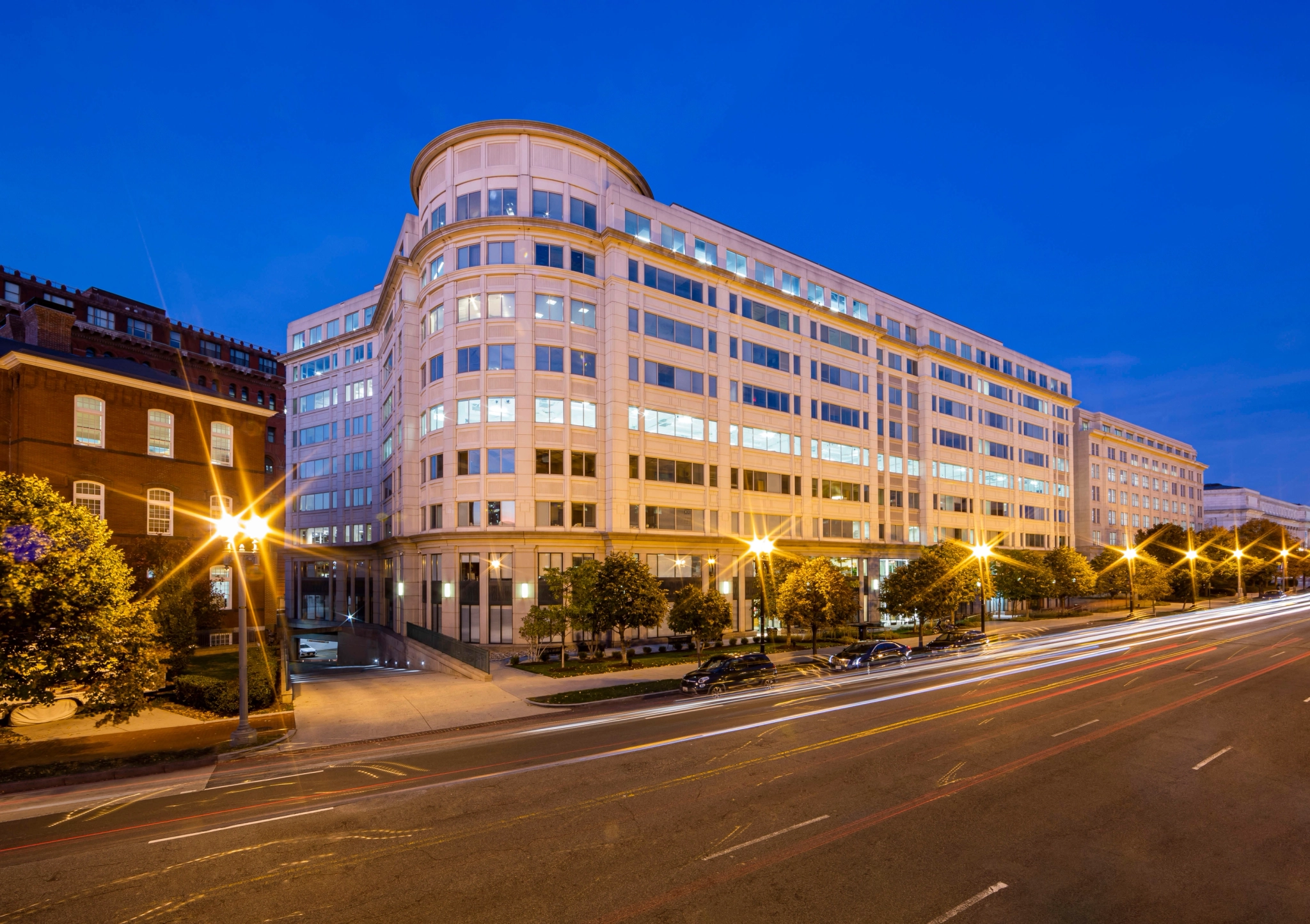 A large modern office building with multiple floors and an exterior of white and glass is illuminated at dusk. Inside, spacious meeting rooms cater to professionals. There are trees and parked cars in front, with light trails from passing vehicles on the street.