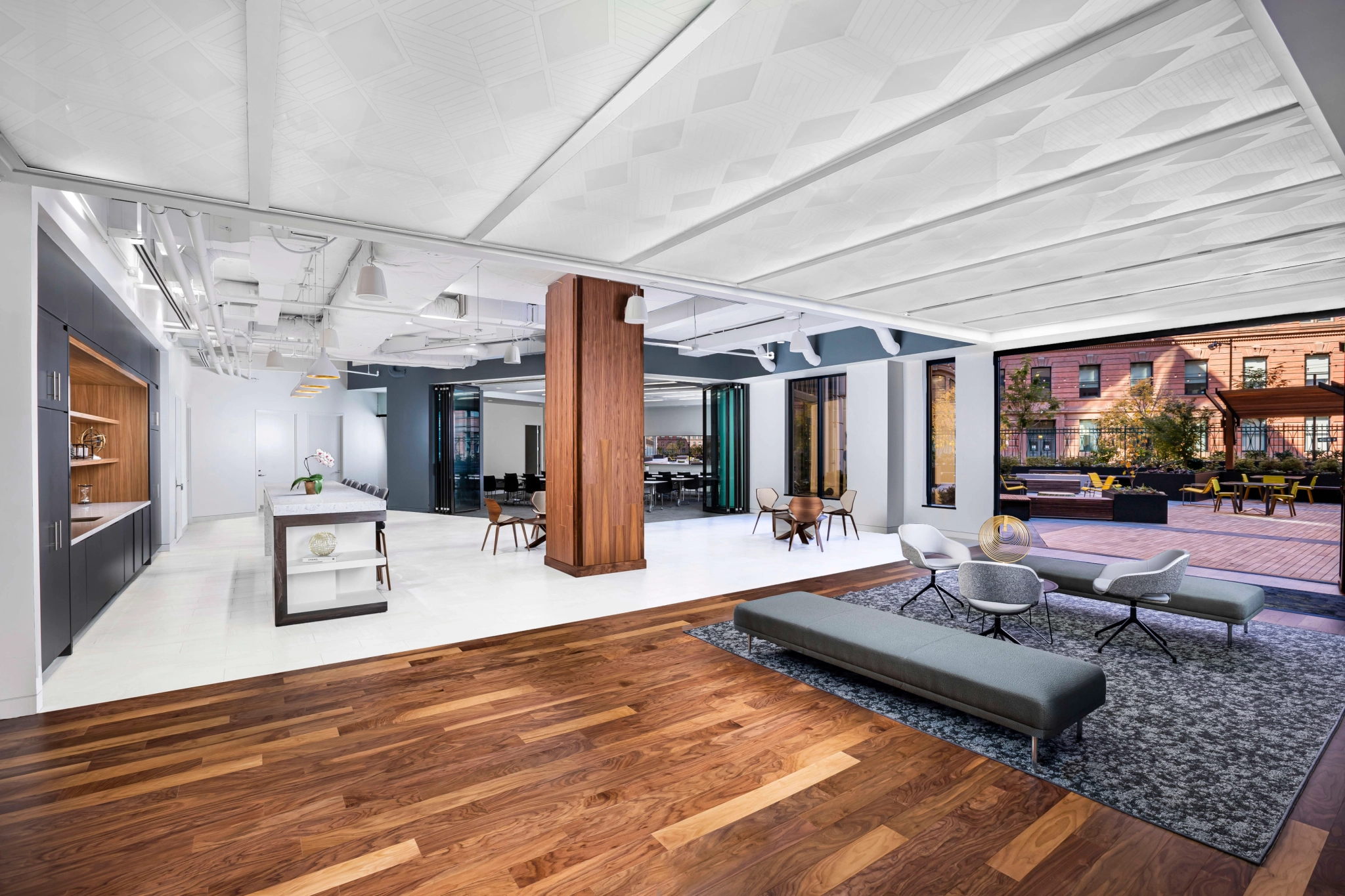 Modern coworking office space with a mix of hardwood and tile flooring, featuring a lounge area, meeting room, kitchenette, and large windows with a view of an outdoor seating area and Washington's urban buildings.