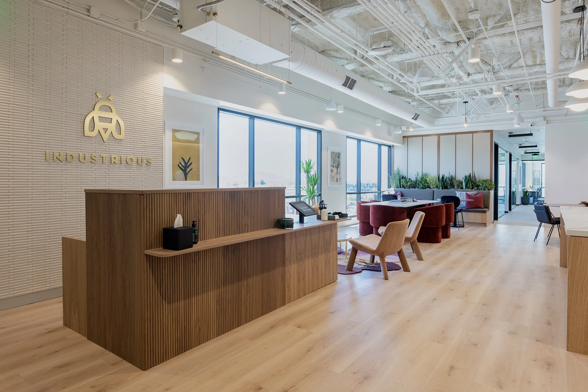 Modern office reception area with wood furnishings, a front desk, seated area with red chairs, large windows, and a sign on the wall reading "INDUSTRIOUS." This inviting workspace also includes access to a nearby meeting room for added convenience.