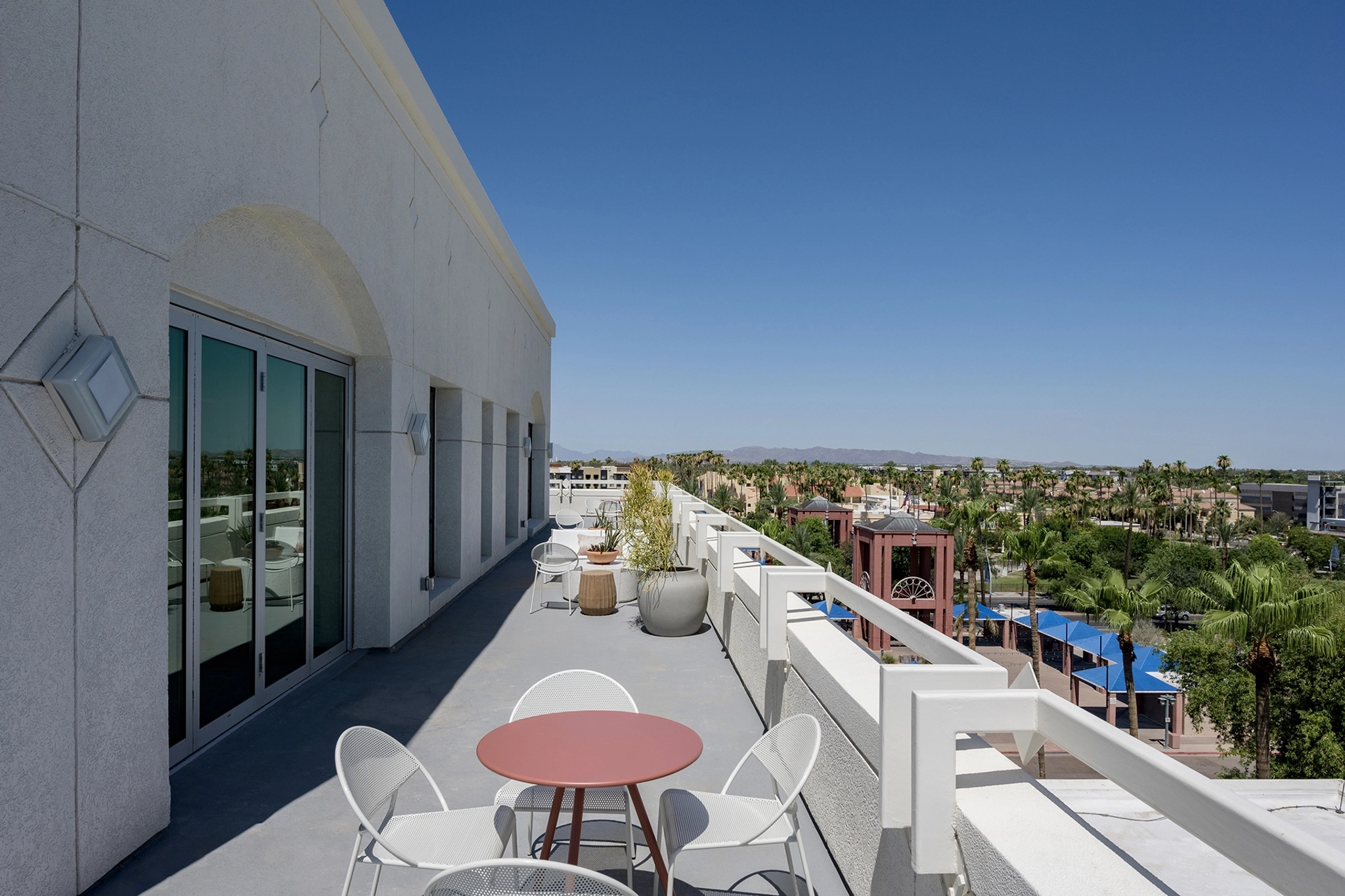 Eine Dachterrasse mit weißen Stühlen, einem rosa runden Tisch, üppigen Pflanzen und Stadtansichten unter einem strahlend blauen Himmel machen dies zum idealen Coworking Space.