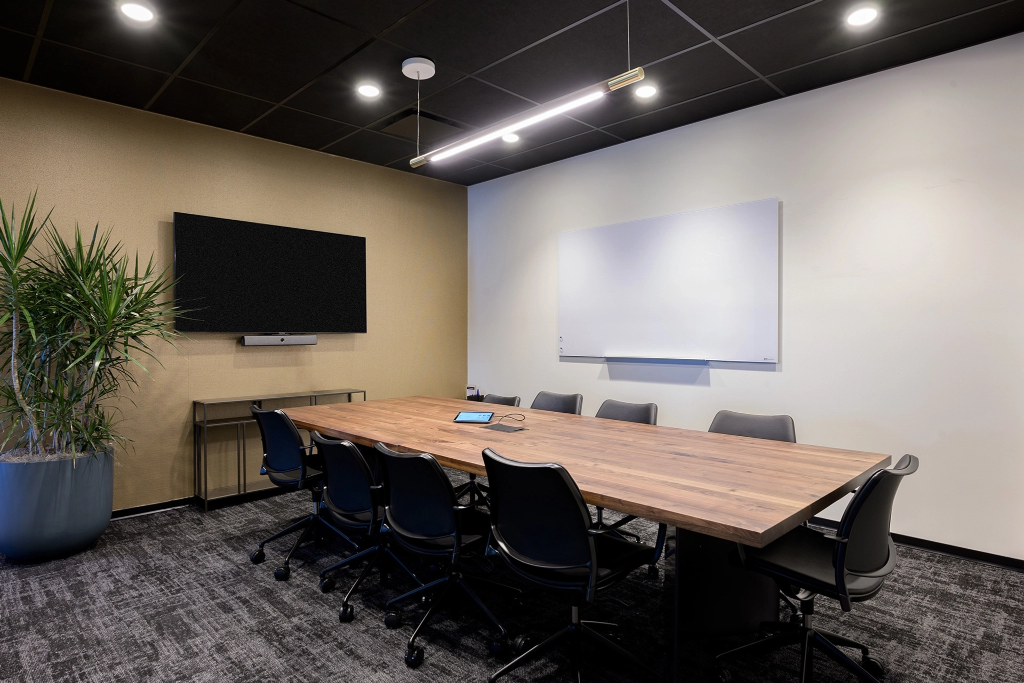 A modern meeting room with a wooden table, eight chairs, a TV screen, a whiteboard, and a potted plant, all set against a backdrop of black carpeted floor and a mix of tan and white walls.