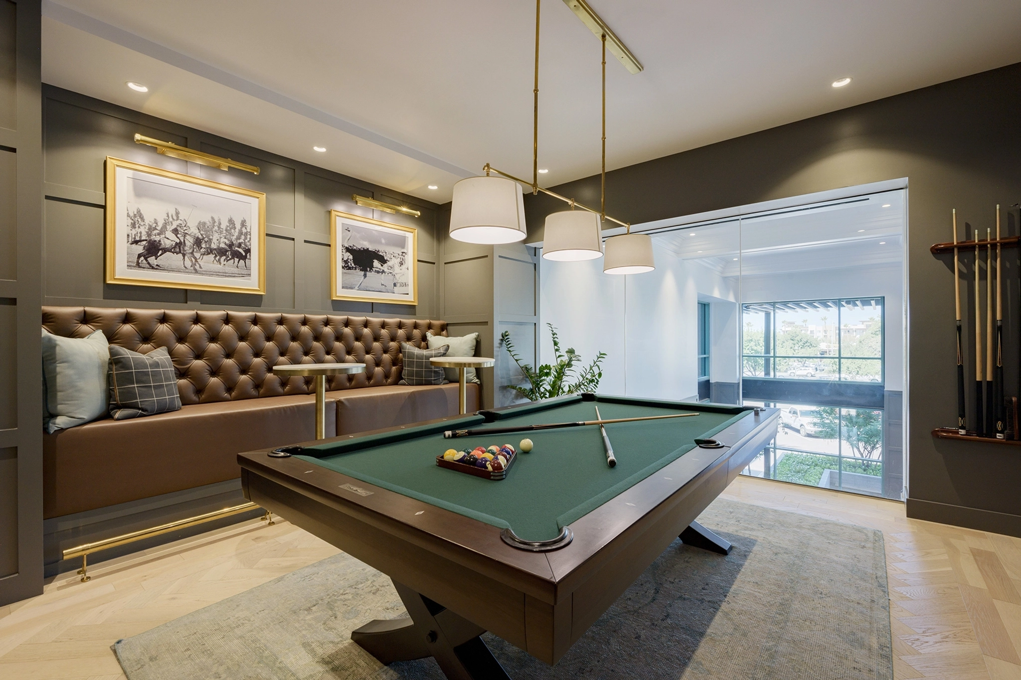 A modern game room with a green pool table, a wall-mounted cue rack, framed pictures on the wall, and a cushioned bench with pillows features glass doors that offer a view of the adjacent coworking space.