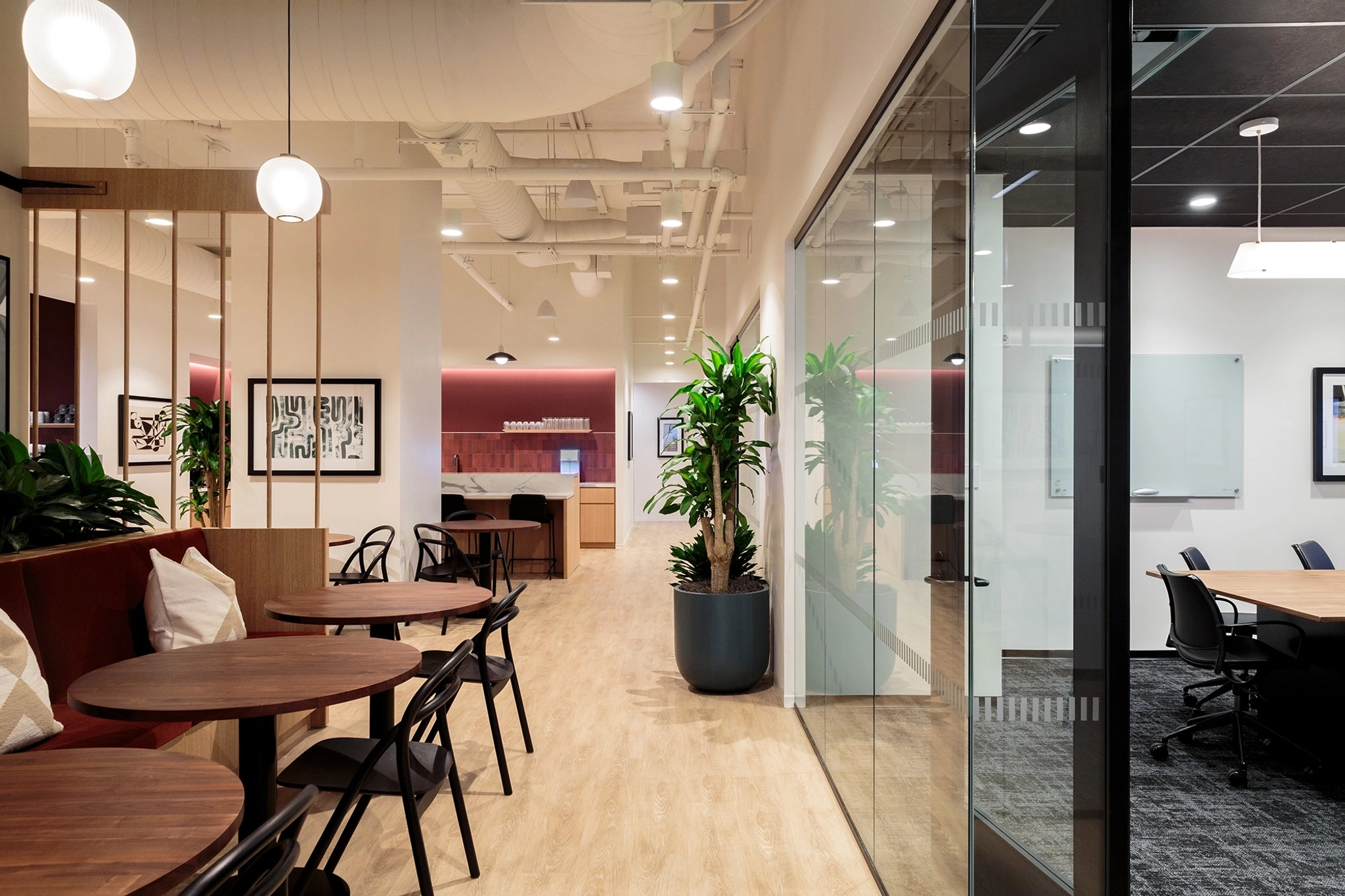 Modern office interior with wooden tables, black chairs, potted plants, and glass partitions. This inviting workspace features bright lighting with ceiling fixtures and artwork adorning the walls.