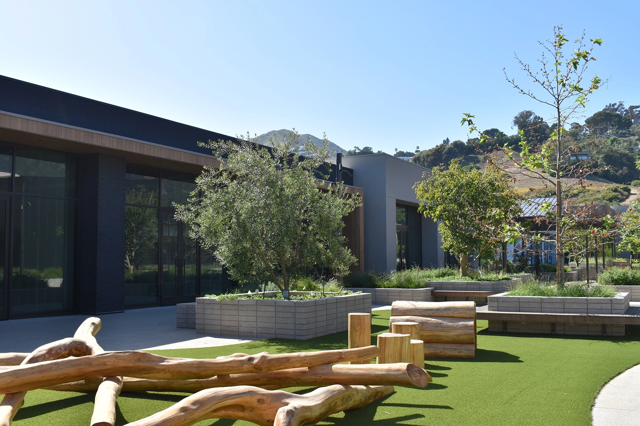 Outdoor courtyard with modern buildings, green artificial turf, trees, and wooden log seating under a clear blue sky—a serene extension of the coworking space.