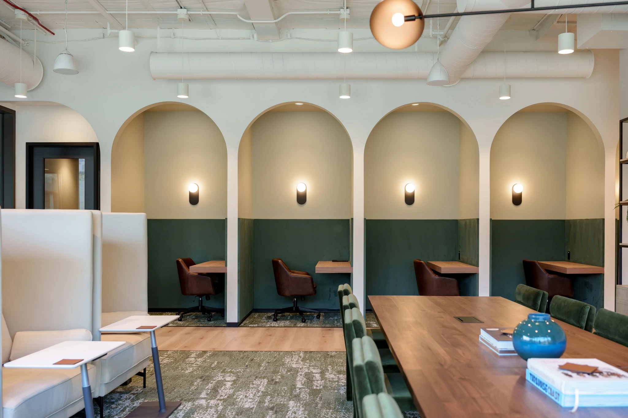 Modern coworking office space with three work nooks under arches, each featuring a chair and desk, soft lighting, and a communal table with books and a vase in the foreground—perfect for those seeking an inspiring Malibu-inspired workspace.