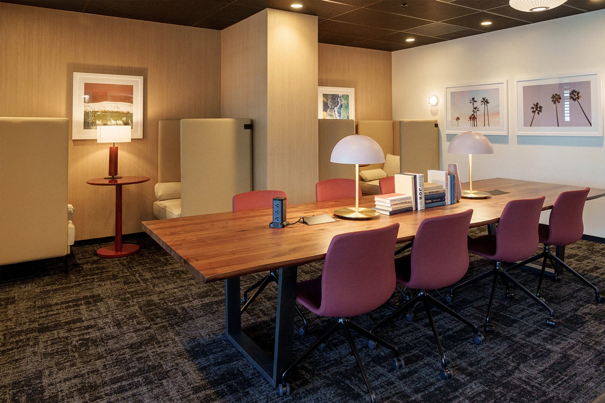 This modern Los Angeles office features a wooden table surrounded by six pink chairs and is accented by two desk lamps. Books and artwork adorn the walls, while beige partitions create semi-private meeting spaces with additional chairs and a small red side table.