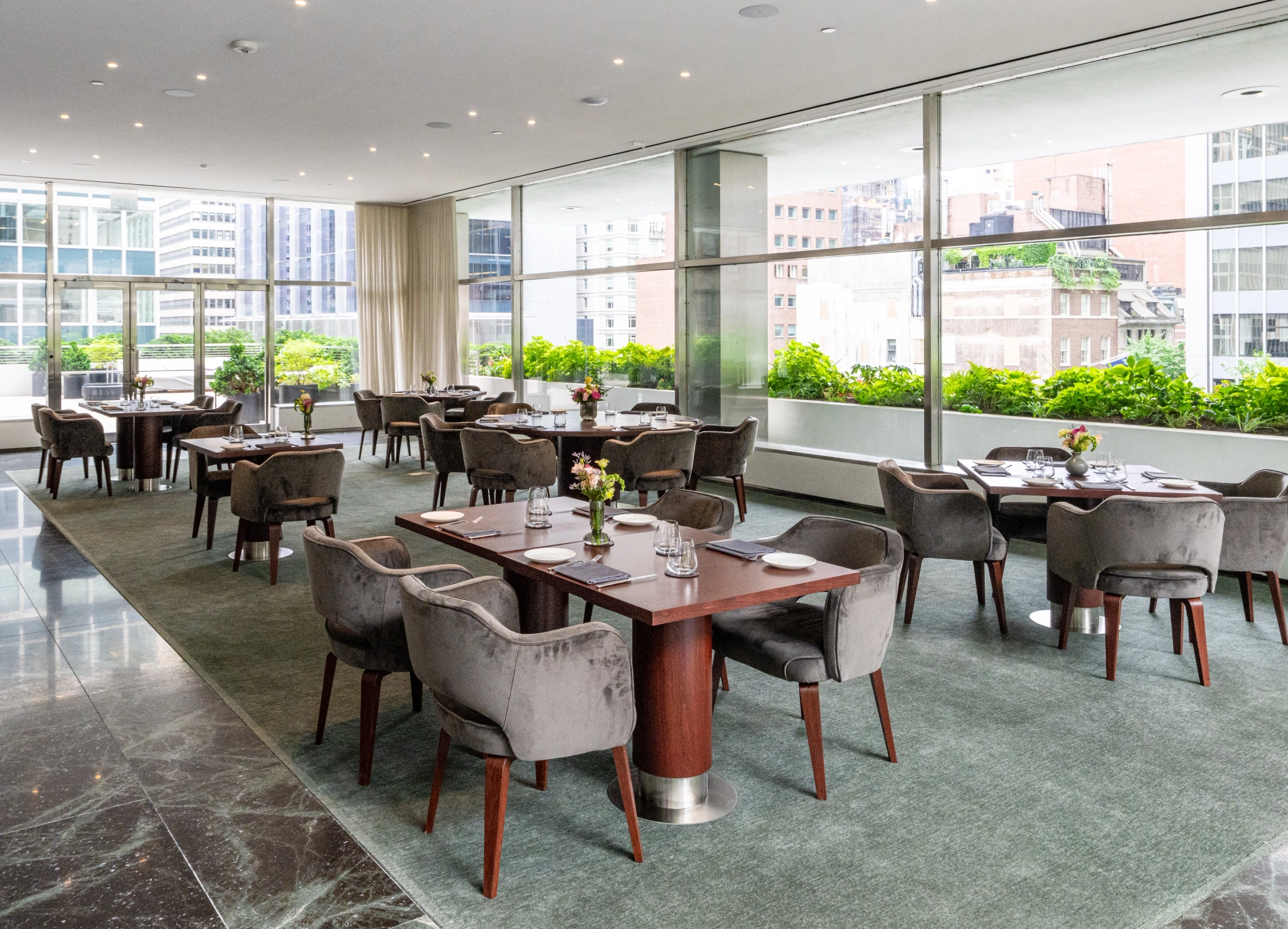 Moderno restaurante con mesas bien organizadas y sillas de terciopelo, con un elegante ambiente de espacio de trabajo y grandes ventanales con vistas a un paisaje urbano con zonas verdes en el exterior.