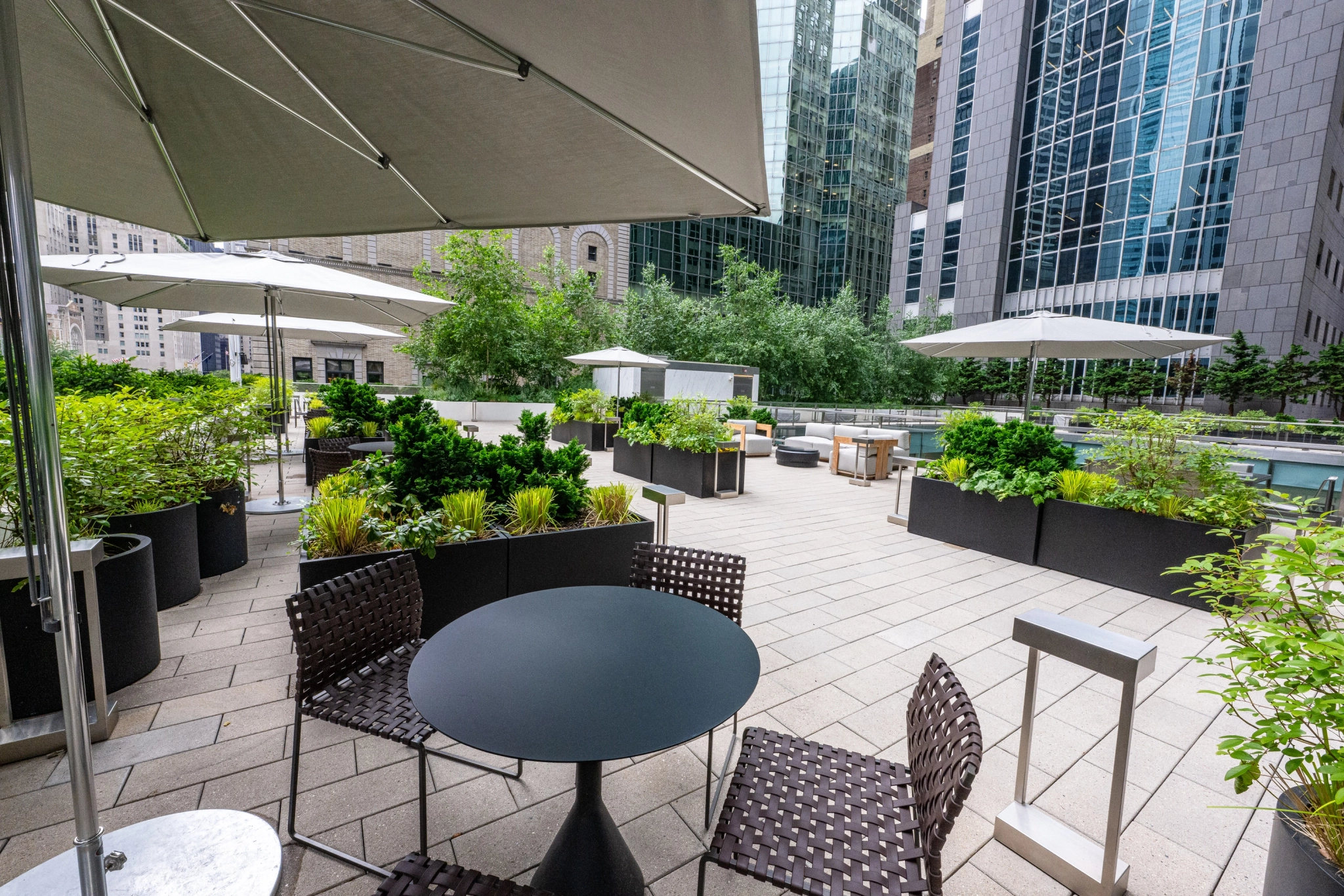 Un elegante espacio de trabajo diseñado como un patio exterior en la azotea con modernas mesas, sillas, plantas en macetas y grandes sombrillas en medio de grandes edificios de oficinas acristalados en un entorno urbano.
