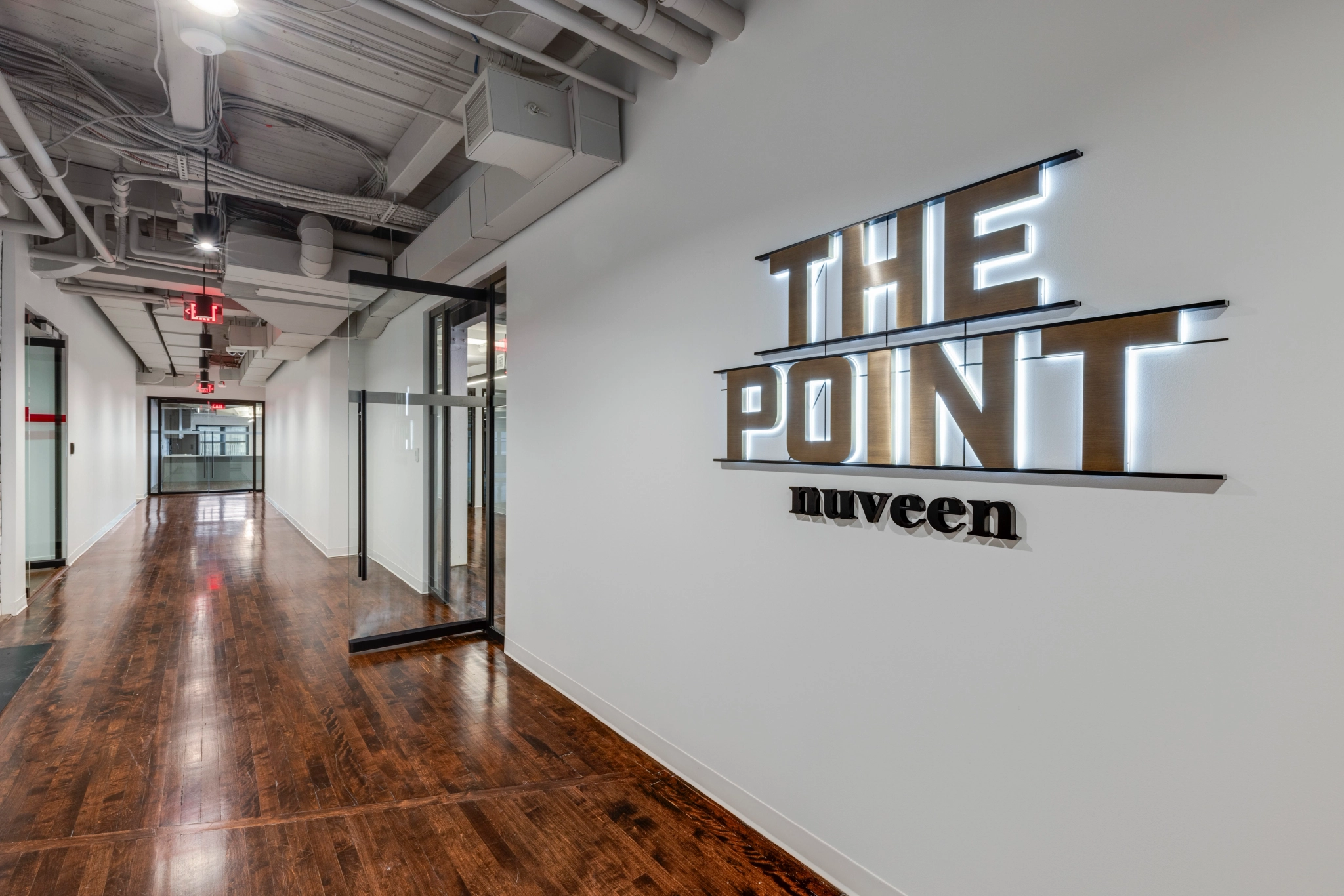 Een gang met houten vloeren en een bord met de tekst “The Point Nuveen” aan de muur leidt naar een moderne vergaderruimte. Op de achtergrond zijn glazen deuren te zien die de bloeiende coworking-scene van Boston weerspiegelen.