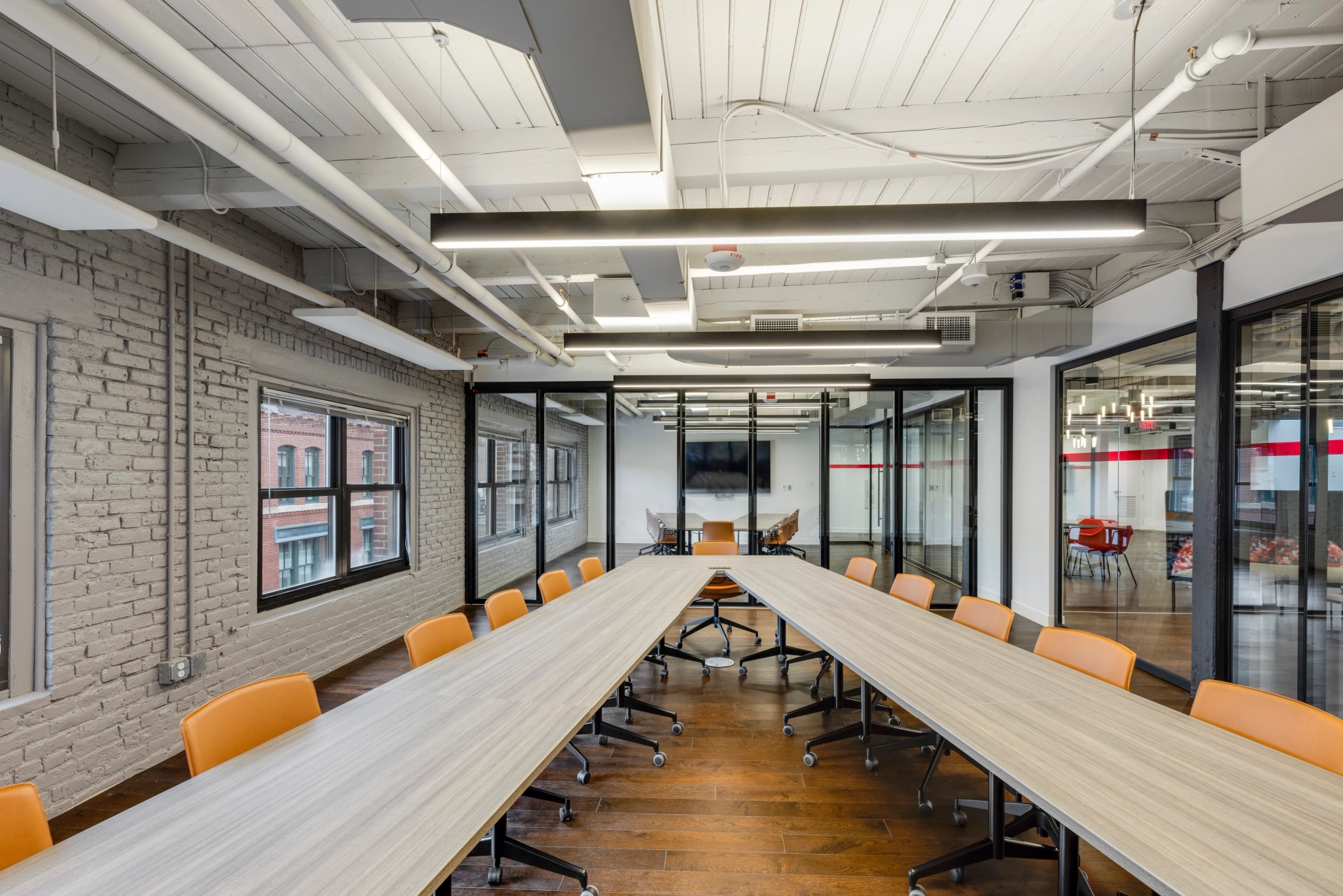 The modern conference room in our Boston coworking space boasts a U-shaped table arrangement with vibrant orange chairs, sleek wood flooring, recessed ceiling lights, and expansive windows for ample natural light.