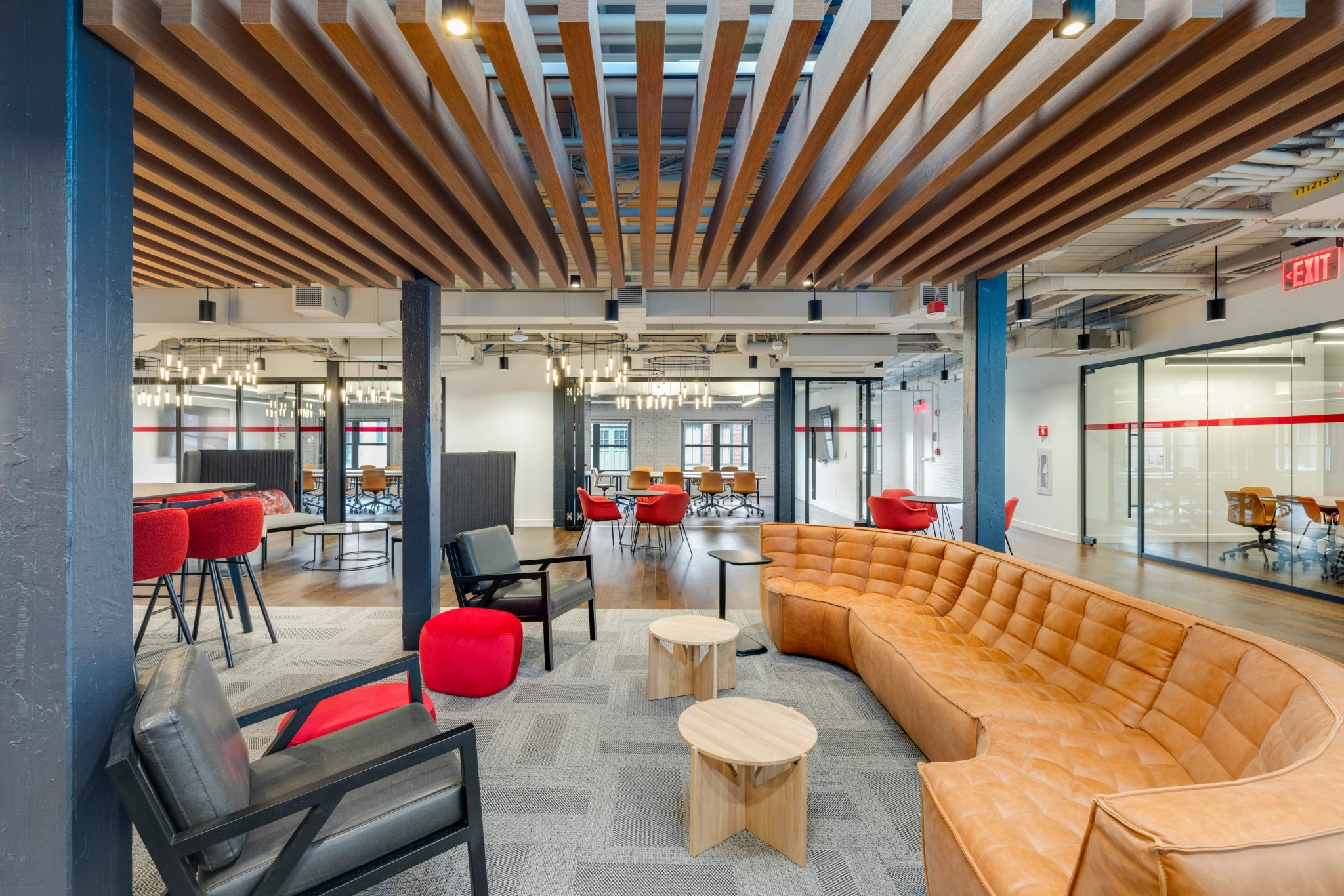 A modern coworking lounge features a brown L-shaped sofa, black and red chairs, round wooden tables, and a ceiling with wooden slats. Glass-walled meeting rooms enhance the collaborative workspace vibe in the background.