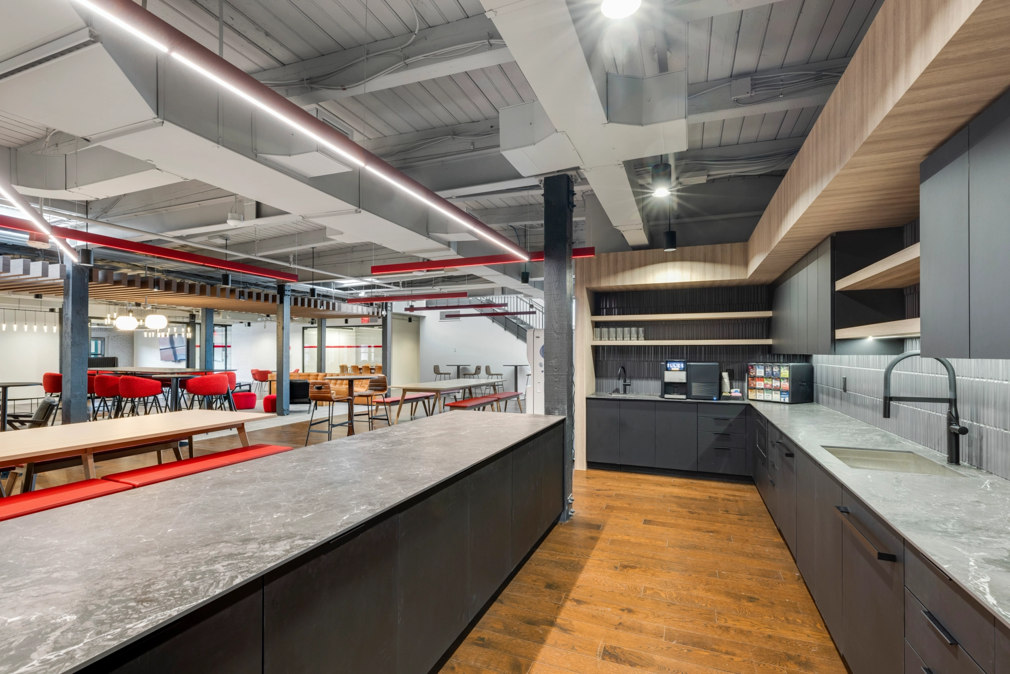 A modern office kitchen features gray cabinets, stone countertops, and wooden flooring. Adjacent to this stylish workspace are red seating and tables in the open area, ideal for informal meetings or casual collaboration.