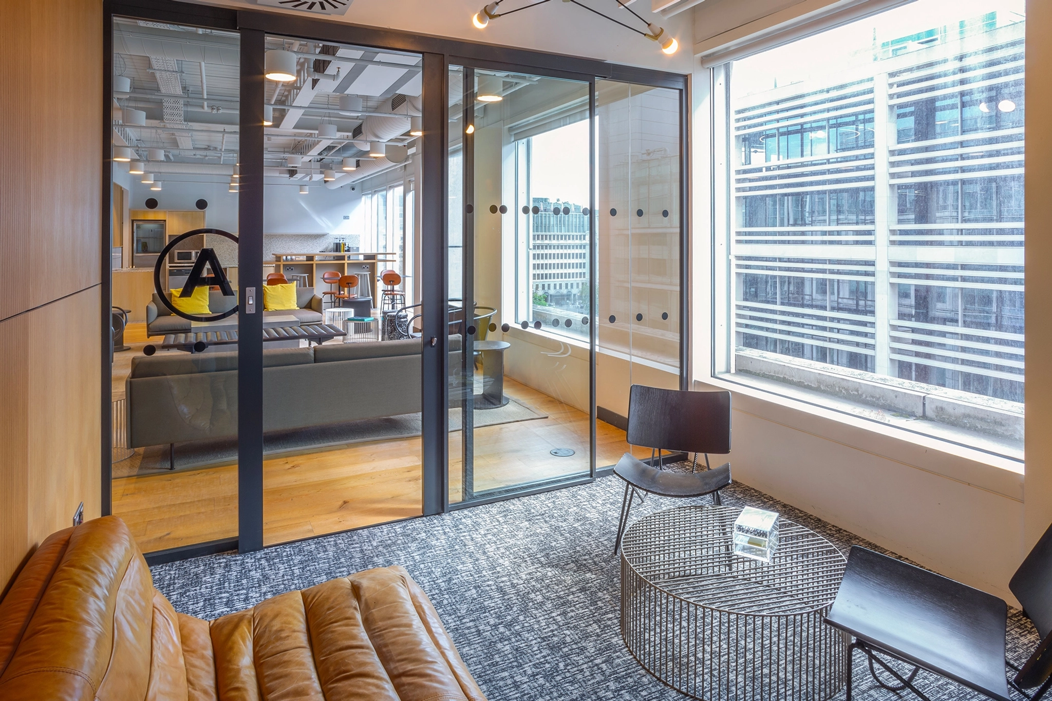 Un espacio de trabajo moderno en Londres con grandes ventanales, paredes de cristal y suelos de madera. Cuenta con sillones de cuero, una mesa de café y vistas a los edificios vecinos, ideal para una elegante sala de reuniones.