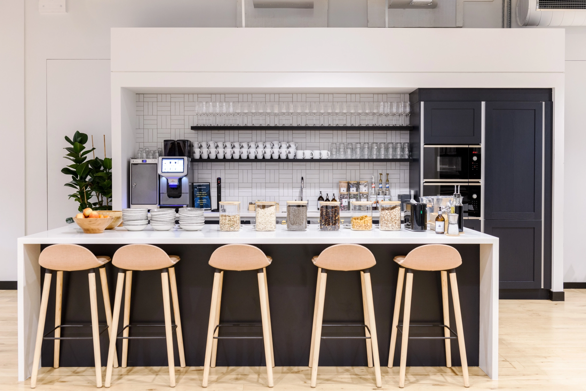 A modern kitchen with a white countertop and four stools, perfect for an informal meeting room vibe. It includes a coffee machine, snack jars, glasses, and plants. Shelves above hold cups and glasses, creating an inviting atmosphere for any London workspace.