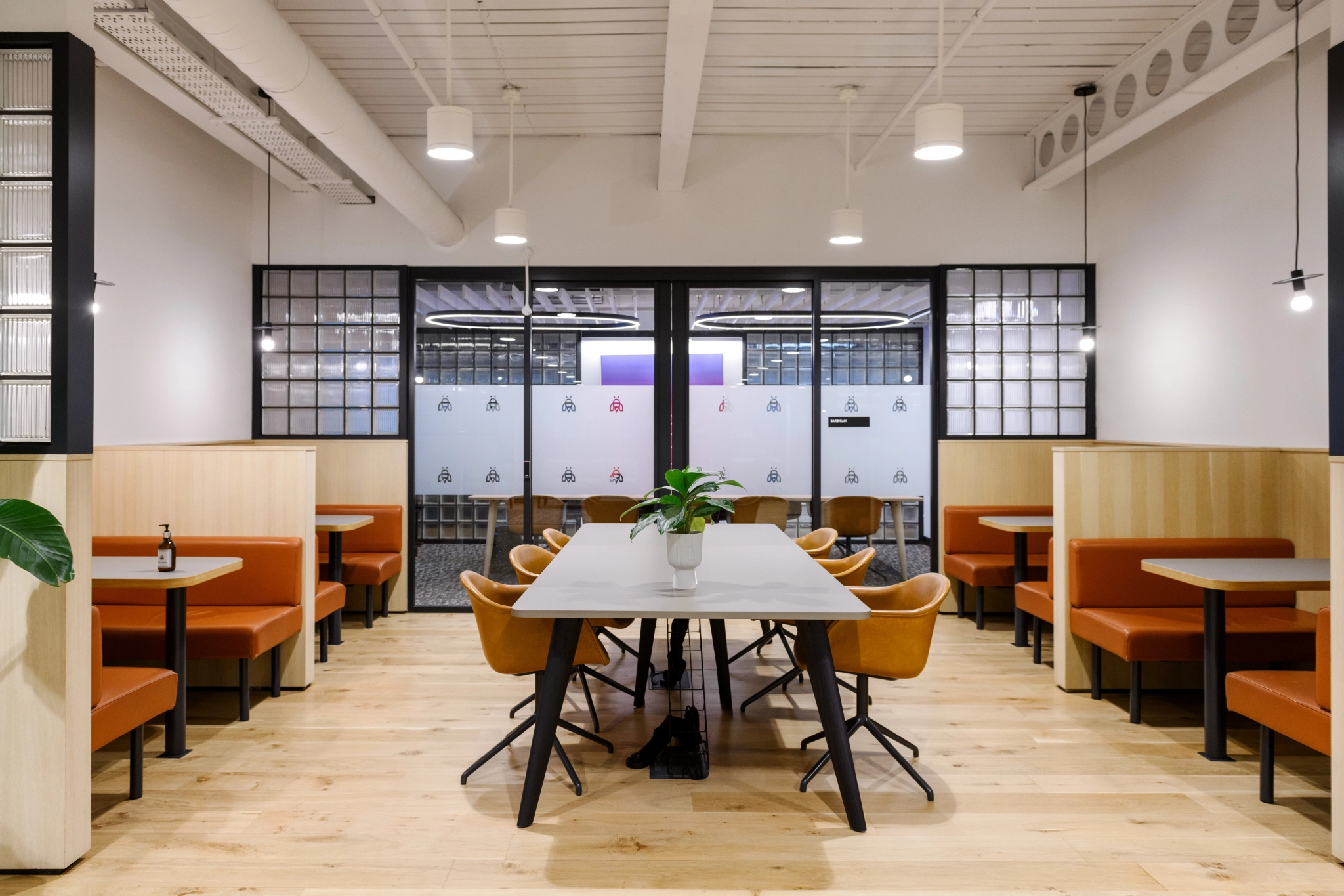 Modern dining area with wooden tables and orange leather chairs, doubling as a stylish coworking space. A potted plant decorates a central table, while glass partitions and industrial ceiling lights complete the professional look.