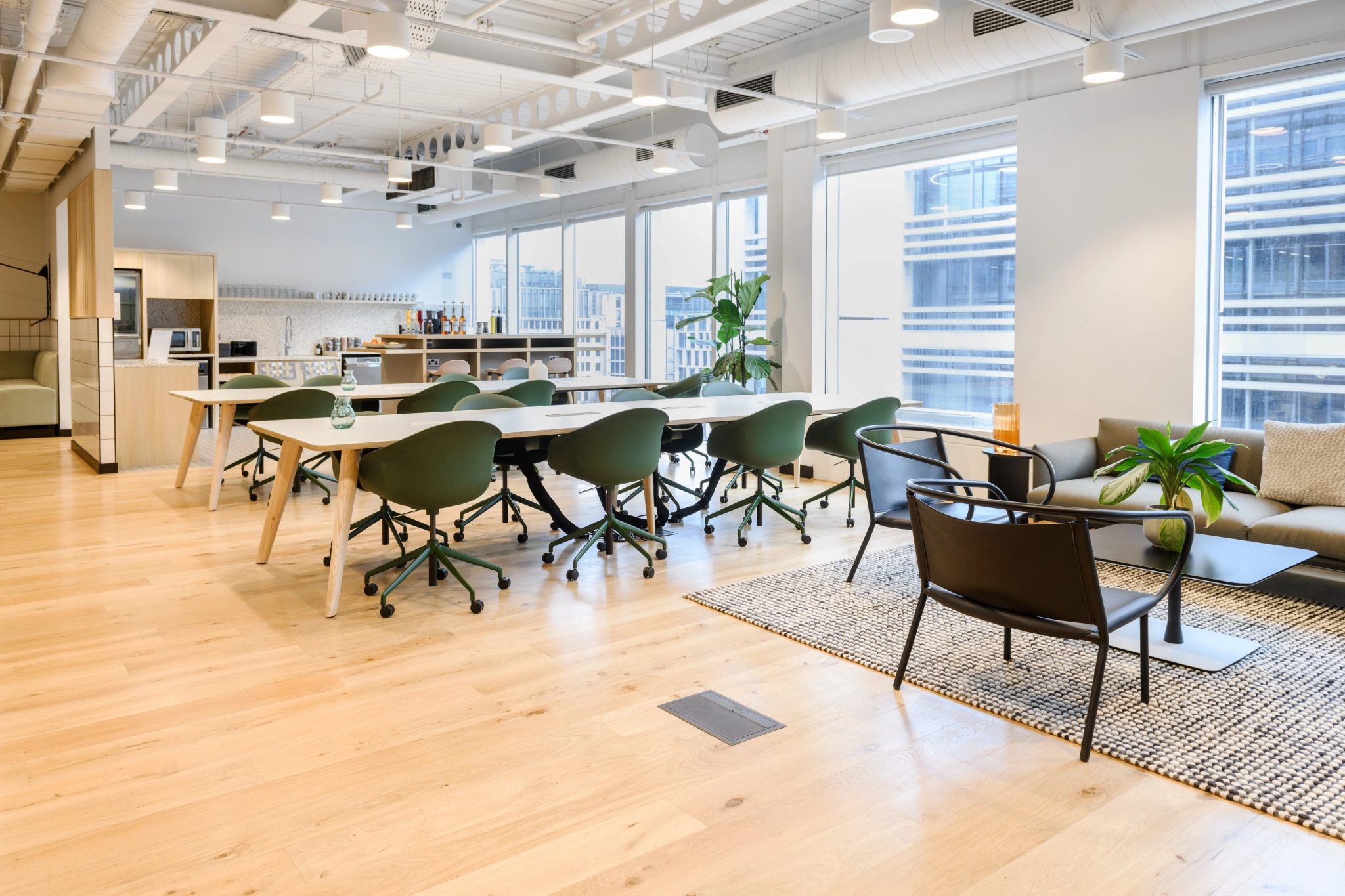 Descubra espacios de oficina modernos en Londres con suelos de madera, paredes blancas y grandes ventanales. Una mesa larga con sillas verdes, junto con un cómodo sofá y sillas, completan la pequeña zona de estar. Es ideal para coworking y está decorado con exuberantes plantas en macetas.