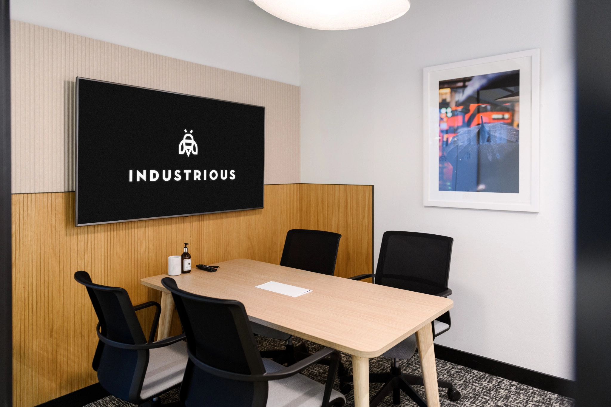 Une salle de réunion élégante et moderne à Londres comprend une table, quatre chaises et un écran mural avec le logo « Industrious ». Une photo encadrée ajoute une touche personnelle à cet espace de travail efficace.