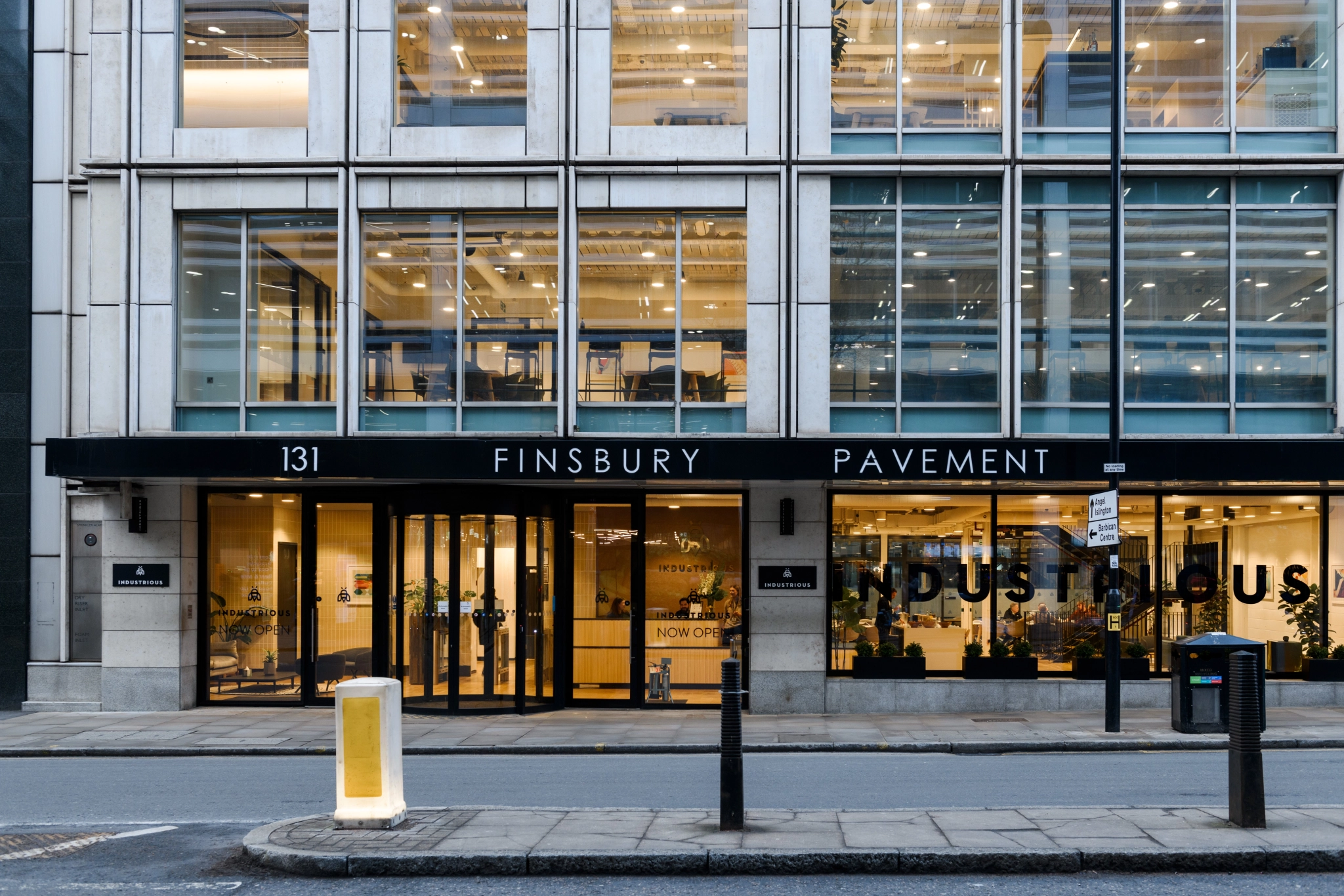 The modern office building facade at 131 Finsbury Pavement, labeled "Industrious," features large windows ideal for coworking in the heart of London. The street in the foreground includes a sign and bollards, enhancing its urban charm.