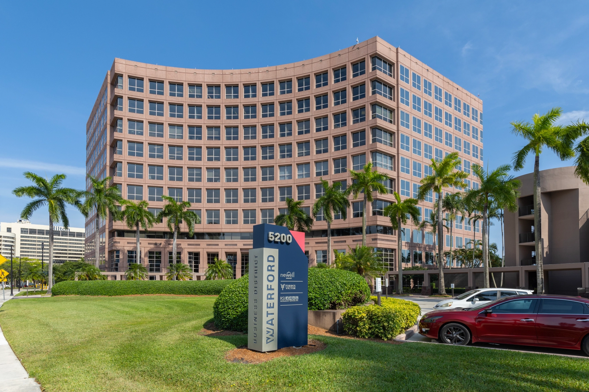 A large, mid-rise office building with a pinkish facade stands under a bright blue sky, surrounded by palm trees. A sign in front reads "WATERFORD 5200," showcasing various business names and coworking spaces.