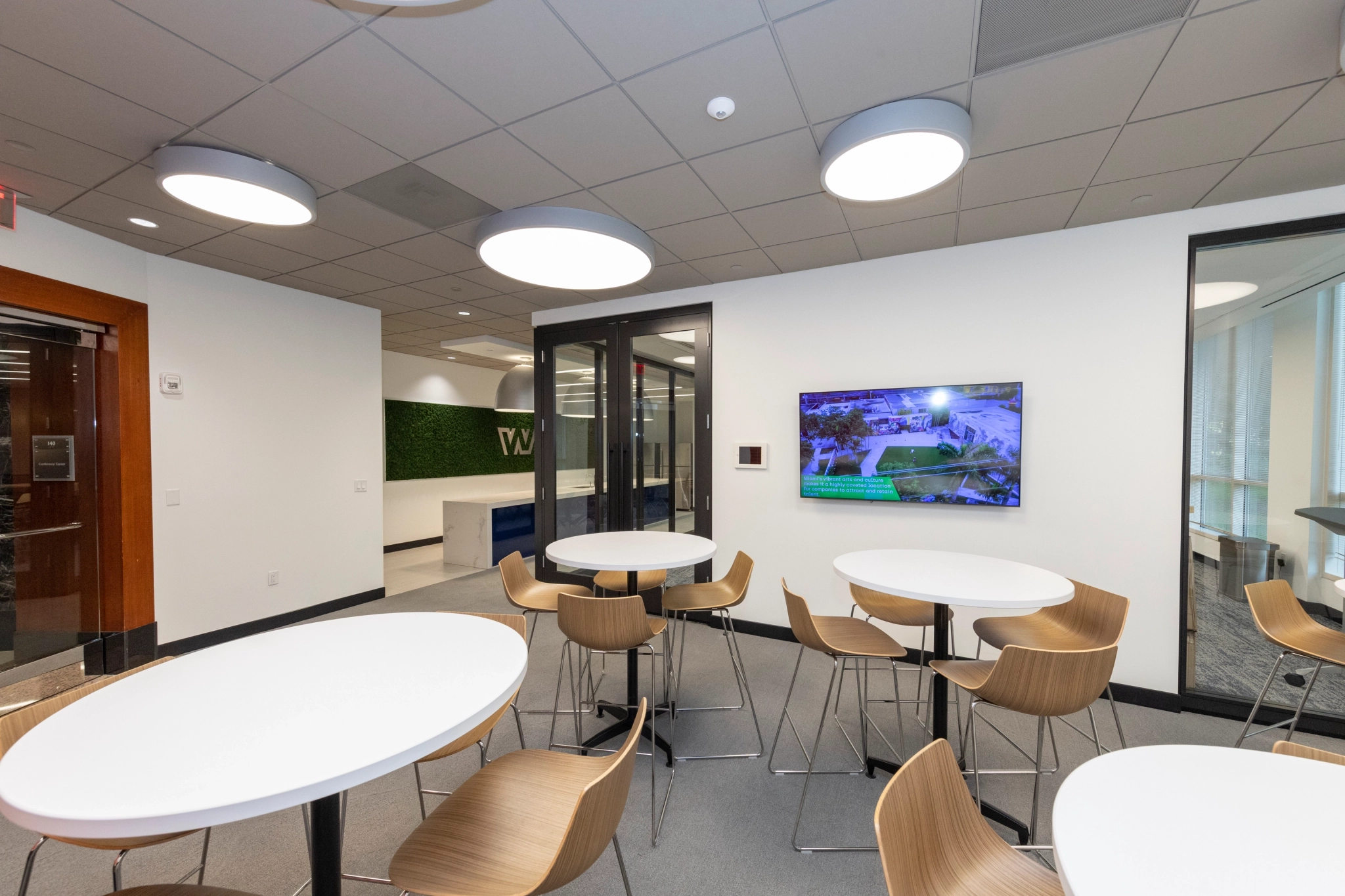 A modern office break area in MIAMI with round white tables, wooden chairs, a wall-mounted TV, and overhead lighting. A hallway leading to the meeting room is visible in the background.