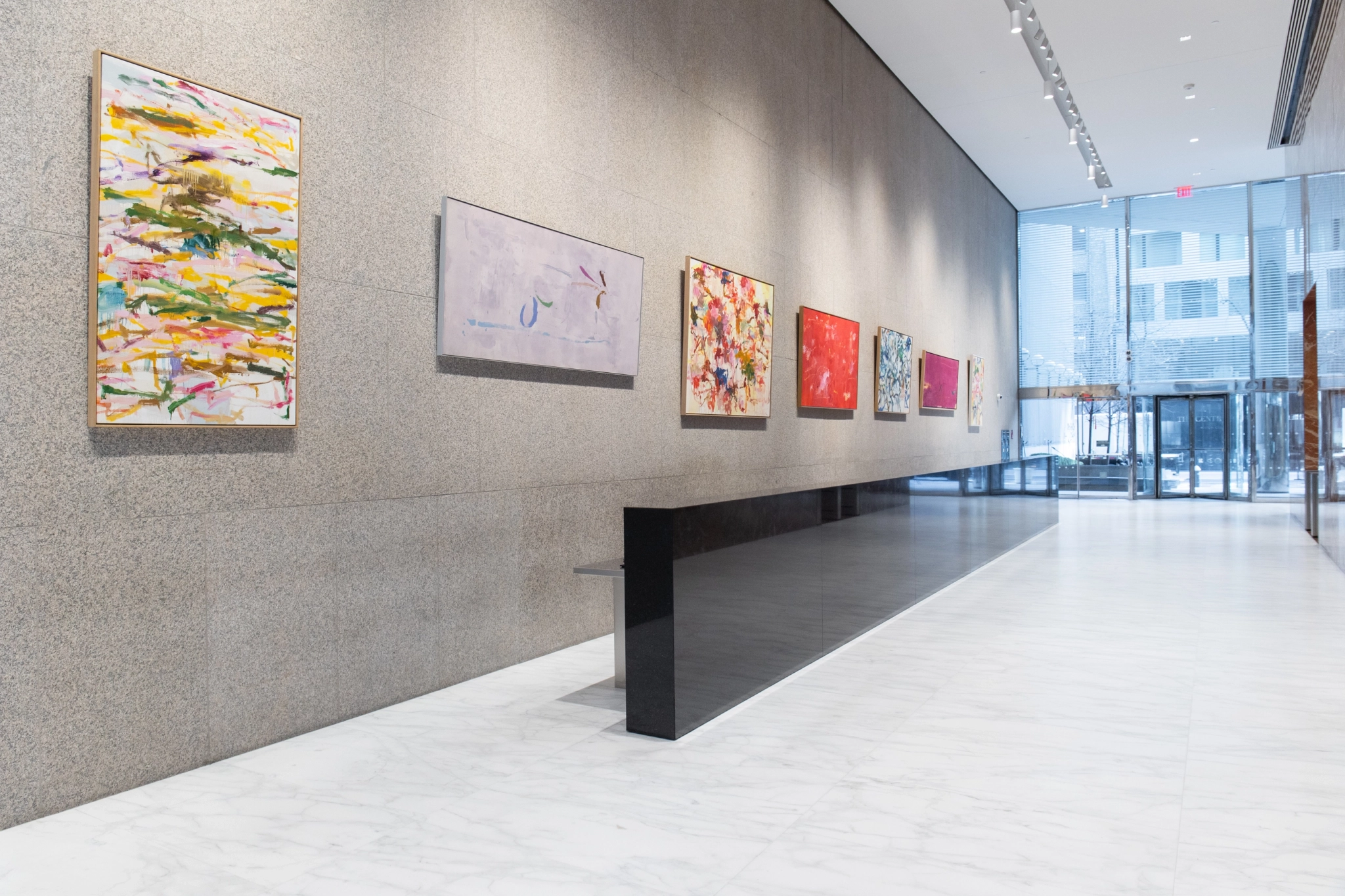 A gallery with a row of abstract paintings displayed on a wall inside a modern building with a marble floor and large windows, akin to an avant-garde coworking space.