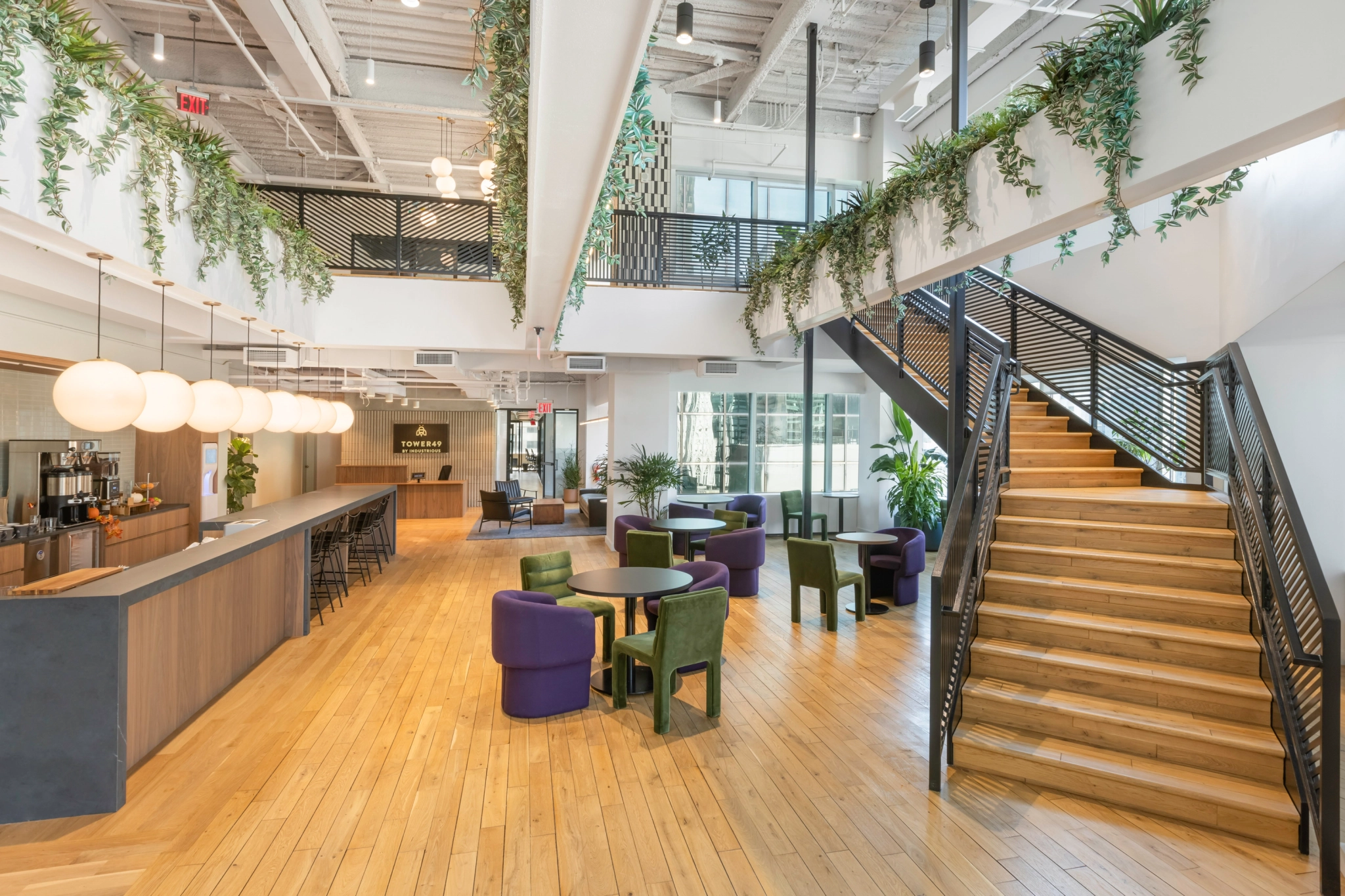 Modern open office space with wooden floors, round tables, purple chairs, lounge seating, a coworking area, staircase, hanging plants, and large windows providing natural light.