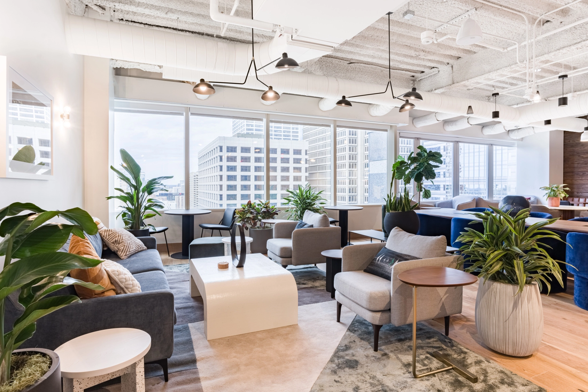 A modern coworking lounge area with various seating options, potted plants, and large windows offering a city view.