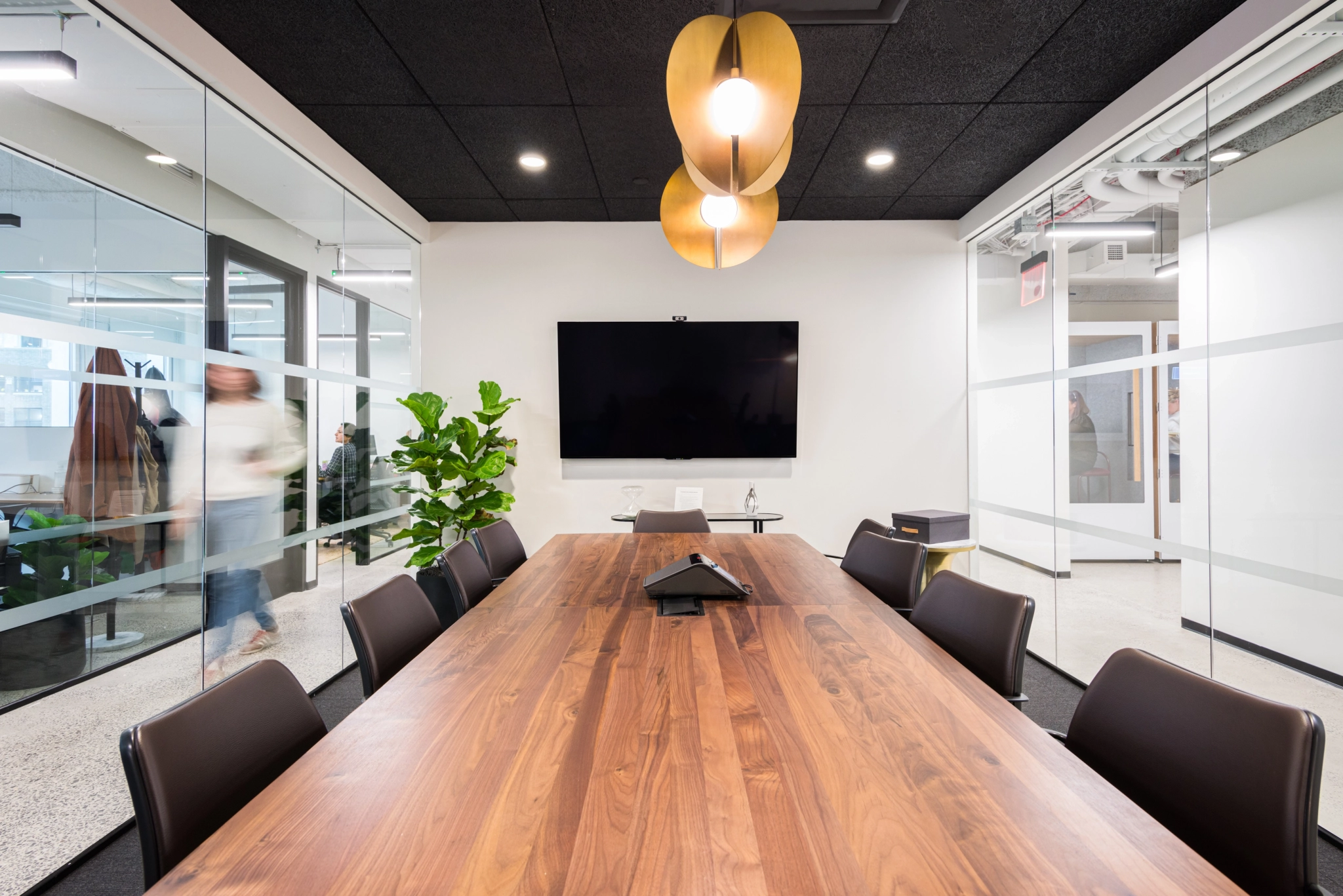 A modern conference room in a bustling New York office features a large wooden table, black chairs, a flat-screen TV on the wall, and glass walls. A person is walking past outside the room in the coworking space.