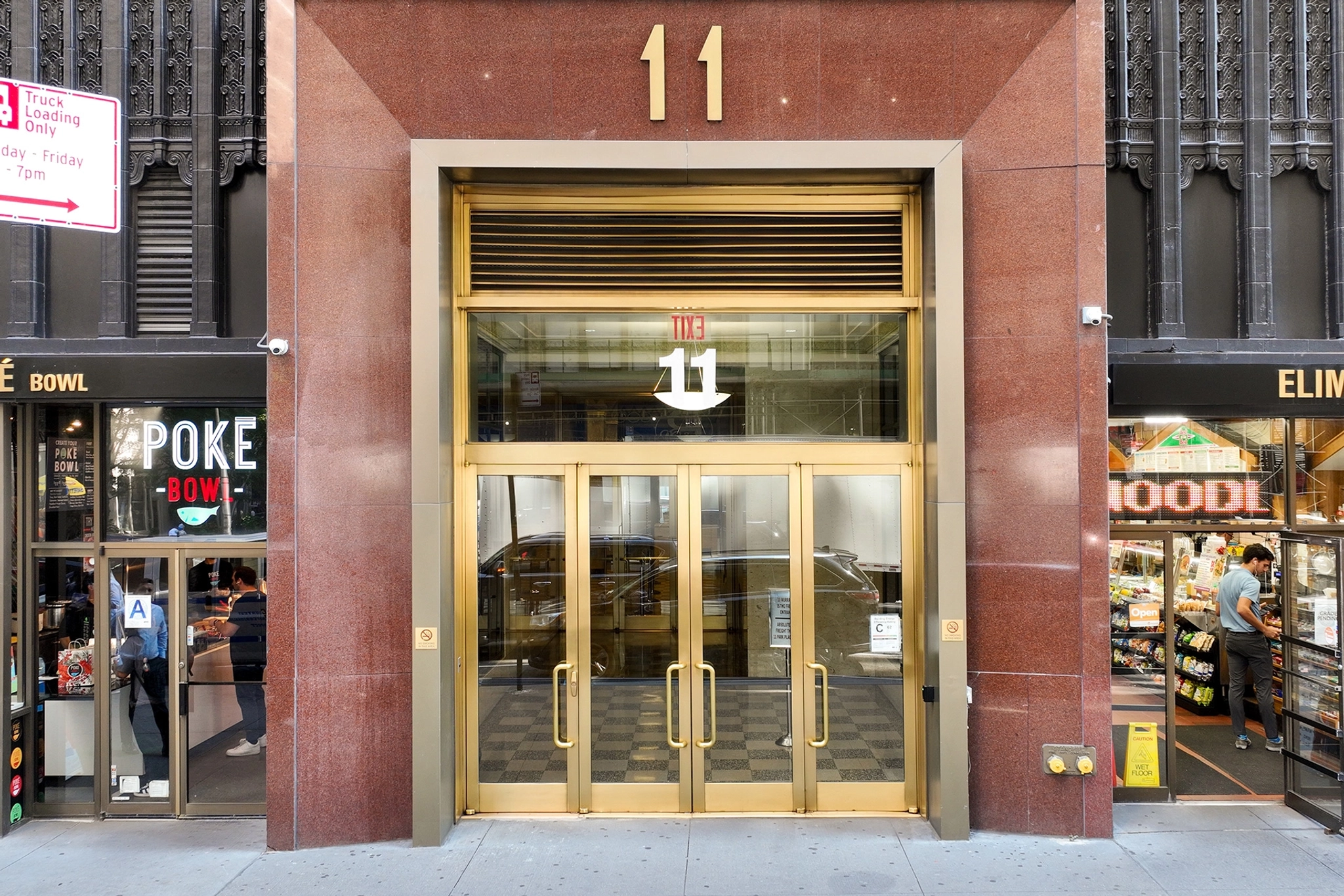 The main entrance of a building with the number 11 above the door, flanked by a poke bowl restaurant on the left and a food store on the right, leads to a bustling coworking office space in New York.