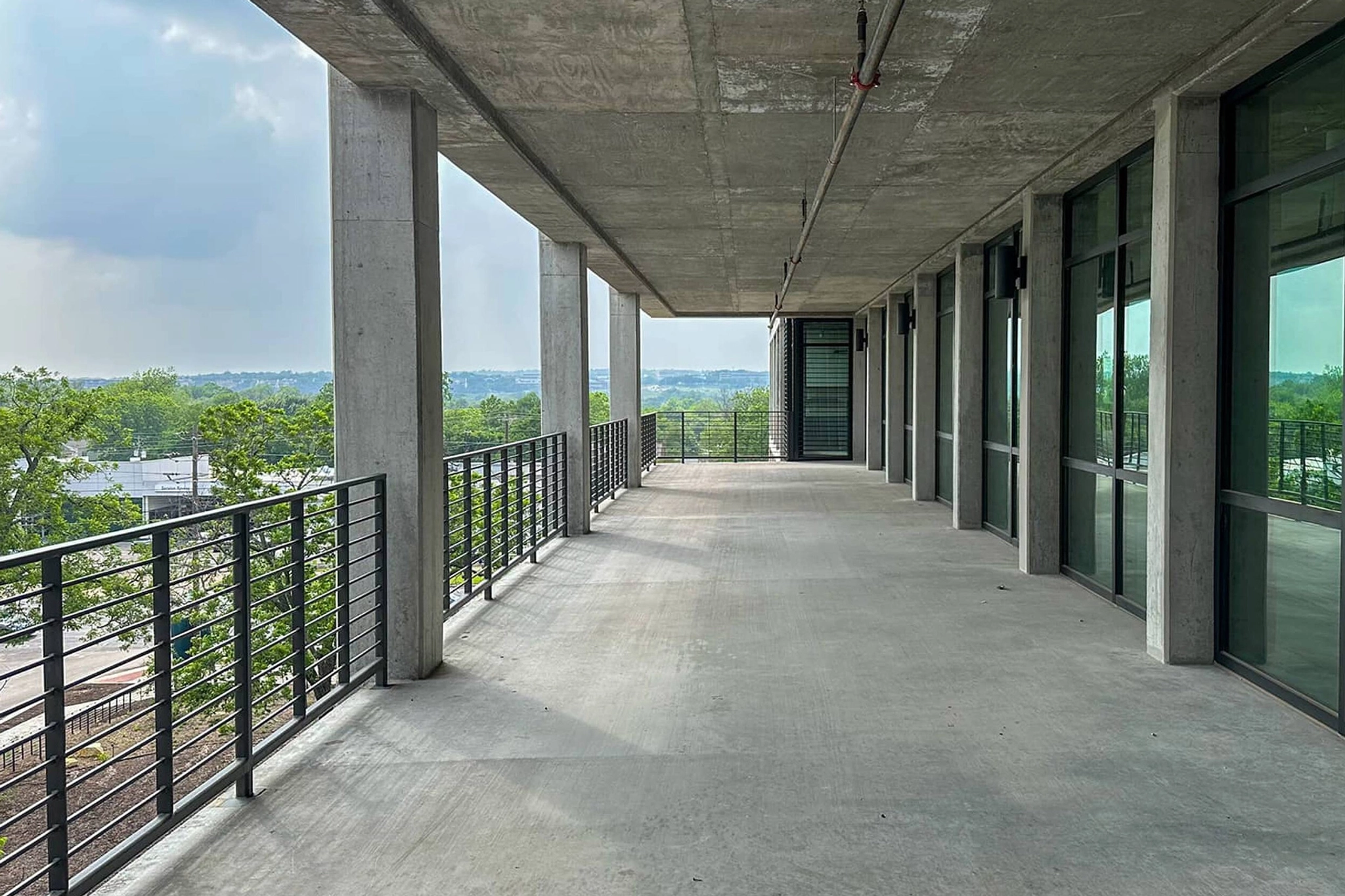 Een groot, leeg betonnen balkon met metalen balustrades en glazen ramen aan één kant kijkt uit over een weelderig, met bomen gevuld landschap onder de bewolkte hemel van Austin en biedt een inspirerende werkruimte.